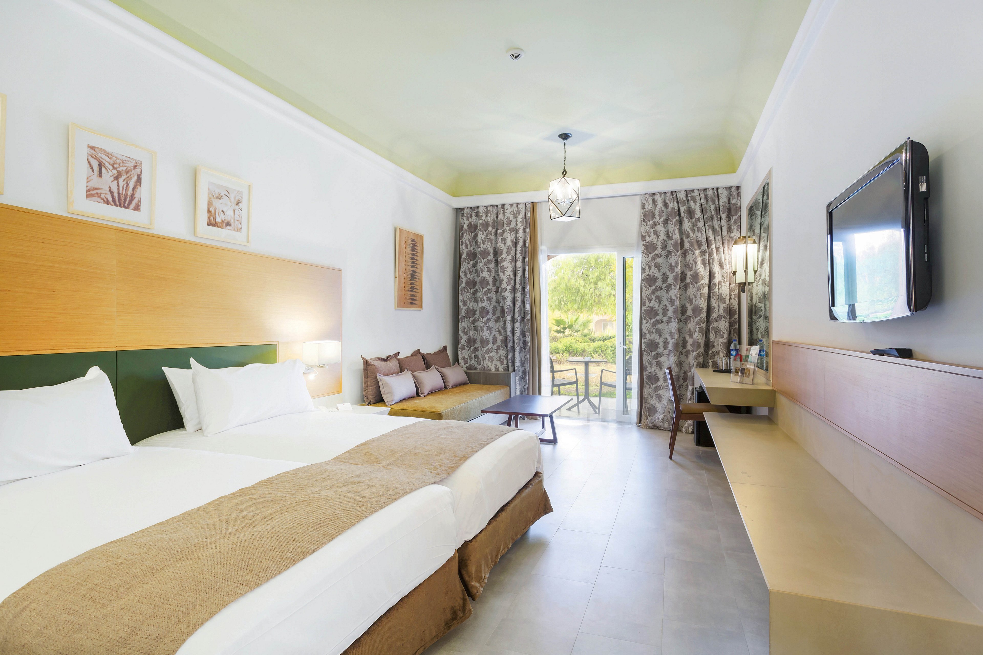 A spacious hotel room features a large double bed with white linens and a beige runner. The room includes a sofa, wall-mounted TV, and a desk, with natural light streaming in from a balcony overlooking greenery. Neutral tones and minimalist decor create a calm, inviting atmosphere, with framed artwork and patterned curtains adding visual interest.