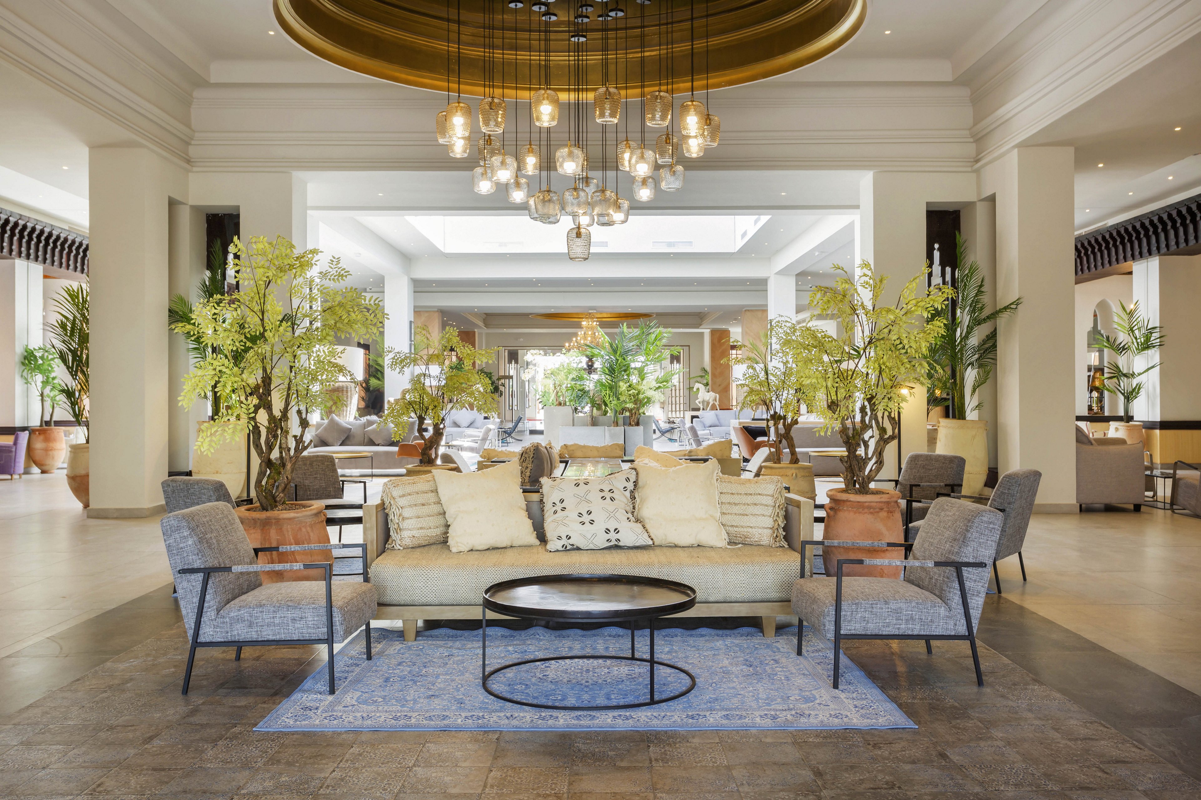 A contemporary hotel lobby features a central seating arrangement with a sofa, armchairs, and a round coffee table atop a blue rug. The space is decorated with lush indoor plants and illuminated by a cluster of hanging pendant lights. Neutral tones and soft textures create a welcoming and elegant atmosphere, with natural light streaming in from large windows in the background.