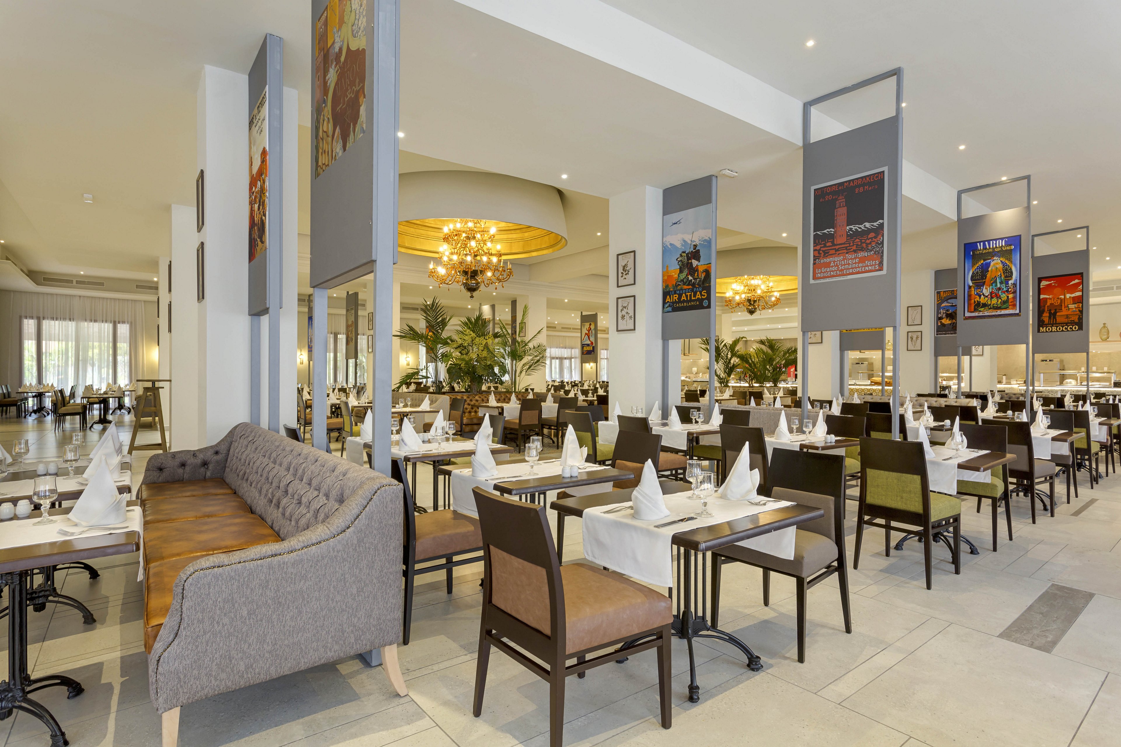The image shows a spacious restaurant with neatly arranged tables and chairs, each set with white napkins and glassware. Large windows allow natural light to fill the room, and decorative posters hang from the ceiling. The setting features neutral tones, contemporary furnishings, and a warm, inviting atmosphere enhanced by a chandelier and indoor plants. No visible text or numeric values are present in the image.