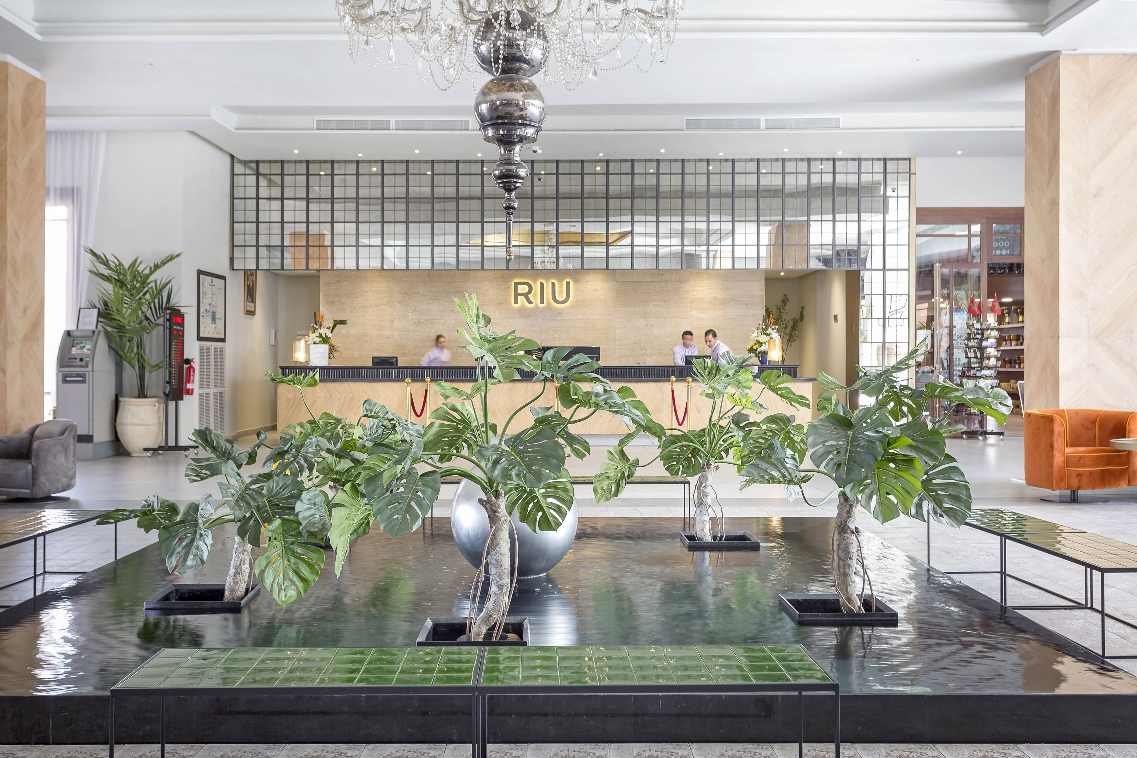 A spacious hotel lobby features a prominent RIU reception desk in the background. The setting includes large green plants in decorative vases, a reflective water feature, and elegant seating areas. The mood is bright and welcoming, with a neutral color palette and contemporary decor. Several people are visible at the reception, and the word 'RIU' is clearly displayed above the desk.