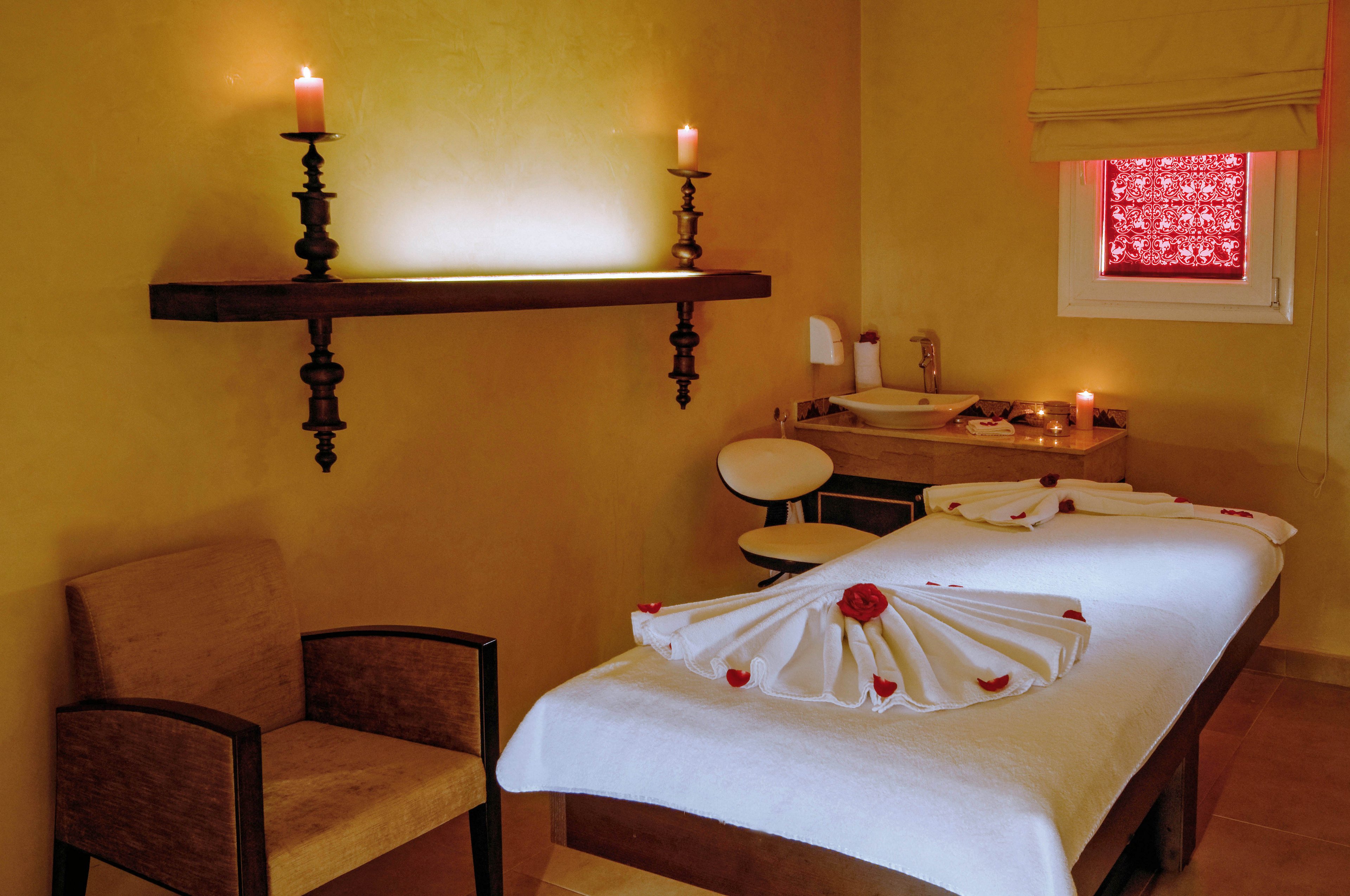 A tranquil spa room features a massage table adorned with neatly folded towels and flower petals. The setting includes warm yellow walls, a shelf with lit candles, and a comfortable chair. Soft lighting and decorative elements create a relaxing ambiance, with a window covered by a patterned shade. No visible text or numeric values are present in the image.