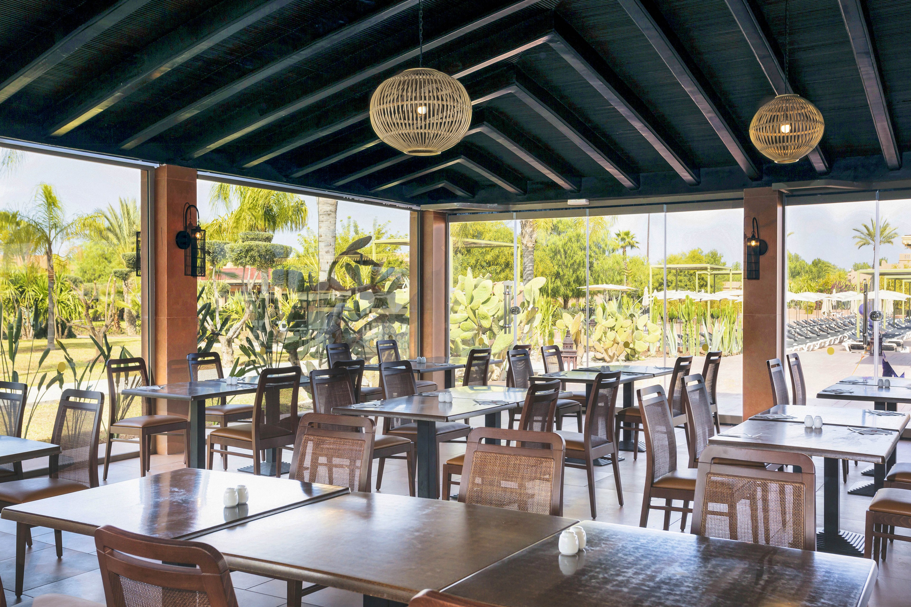 A spacious, contemporary restaurant features wooden tables and chairs arranged neatly under a dark, slanted roof. Large glass windows provide a clear view of lush greenery and palm trees outside, creating a relaxed and airy atmosphere. Natural light fills the space, highlighting the minimalist decor and woven pendant lights. No visible people or text are present in the image.