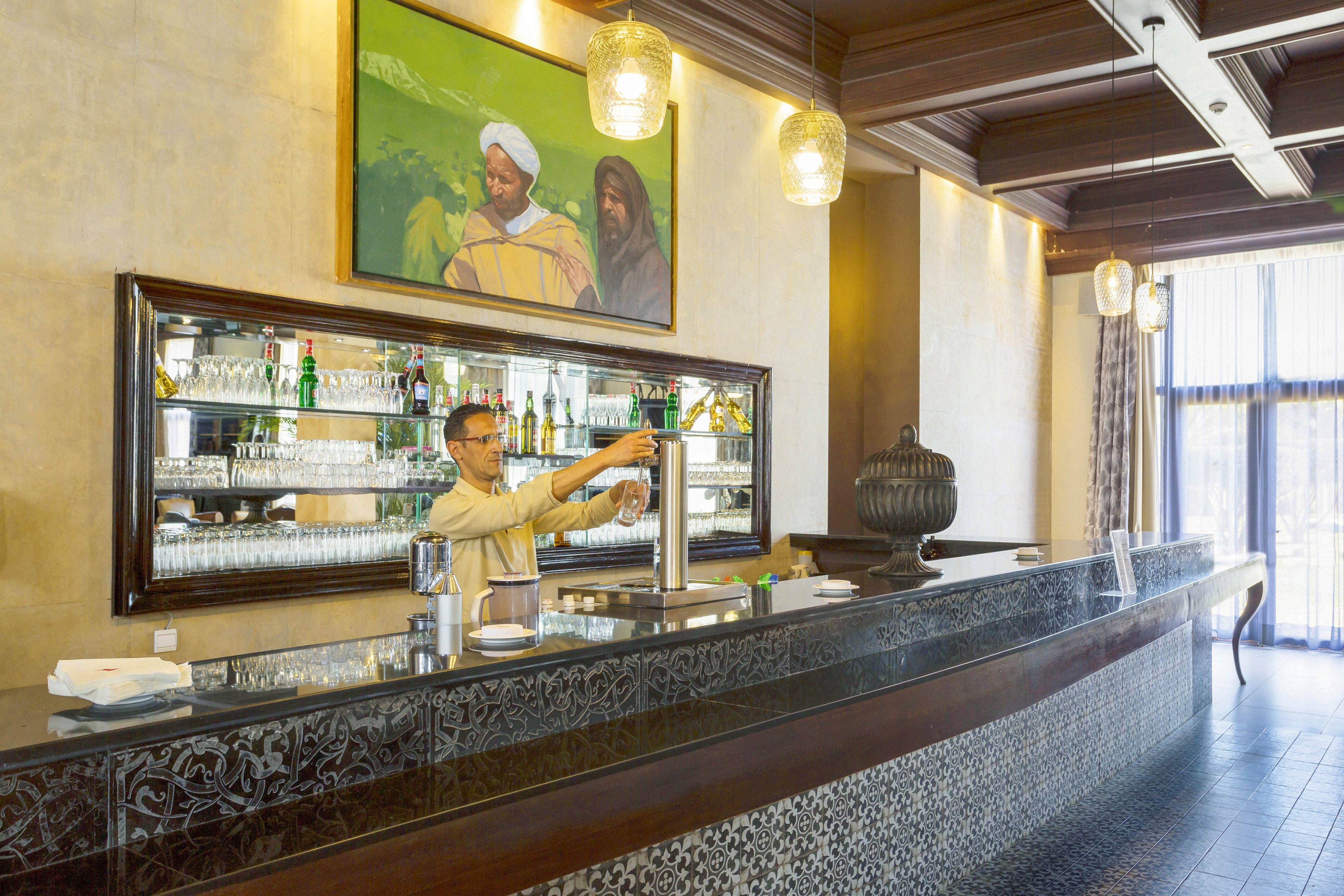 A bartender stands behind a sleek, modern bar counter in a well-lit hotel lounge. The setting features glass shelves with bottles and glassware, decorative hanging lights, and a large window allowing natural light. The bartender appears to be preparing a drink, adding to the lively yet relaxed atmosphere. A contemporary painting and intricate tilework add visual interest to the scene.
