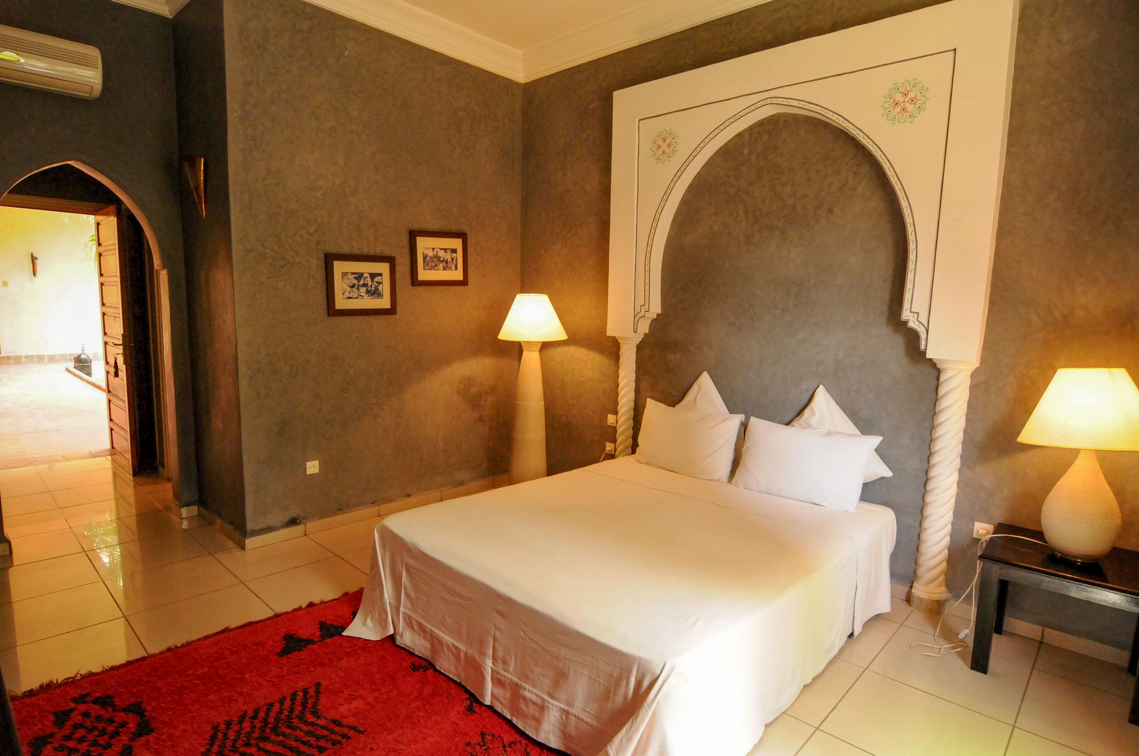 A neatly made double bed with white linens sits against an ornate arched wall feature in a warmly lit bedroom. Two bedside tables hold modern lamps that cast a soft glow on the textured gray walls. A bright red rug adds color to the tiled floor, while framed artwork decorates the wall. An open doorway reveals a glimpse of an adjacent hallway, enhancing the sense of space and comfort.