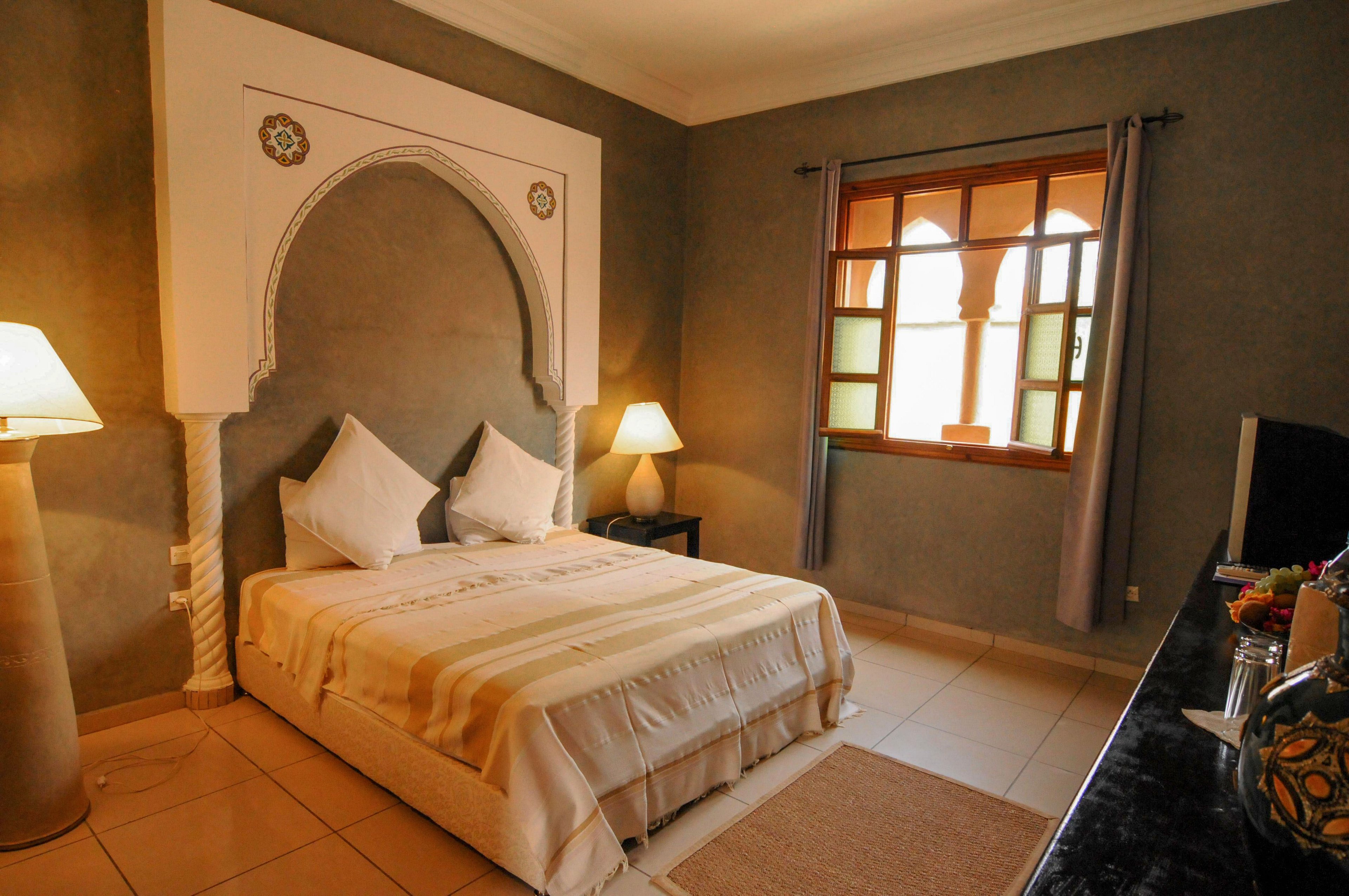 A warm, inviting bedroom features a double bed framed by an ornate Moroccan-style arch. Soft lighting from a bedside lamp and natural light from a wooden window create a comfortable atmosphere. Neutral tones in the bedding, curtains, and tiled floor contribute to a calm, minimalist style. Decorative pillows and subtle wall details add character to the interior space.