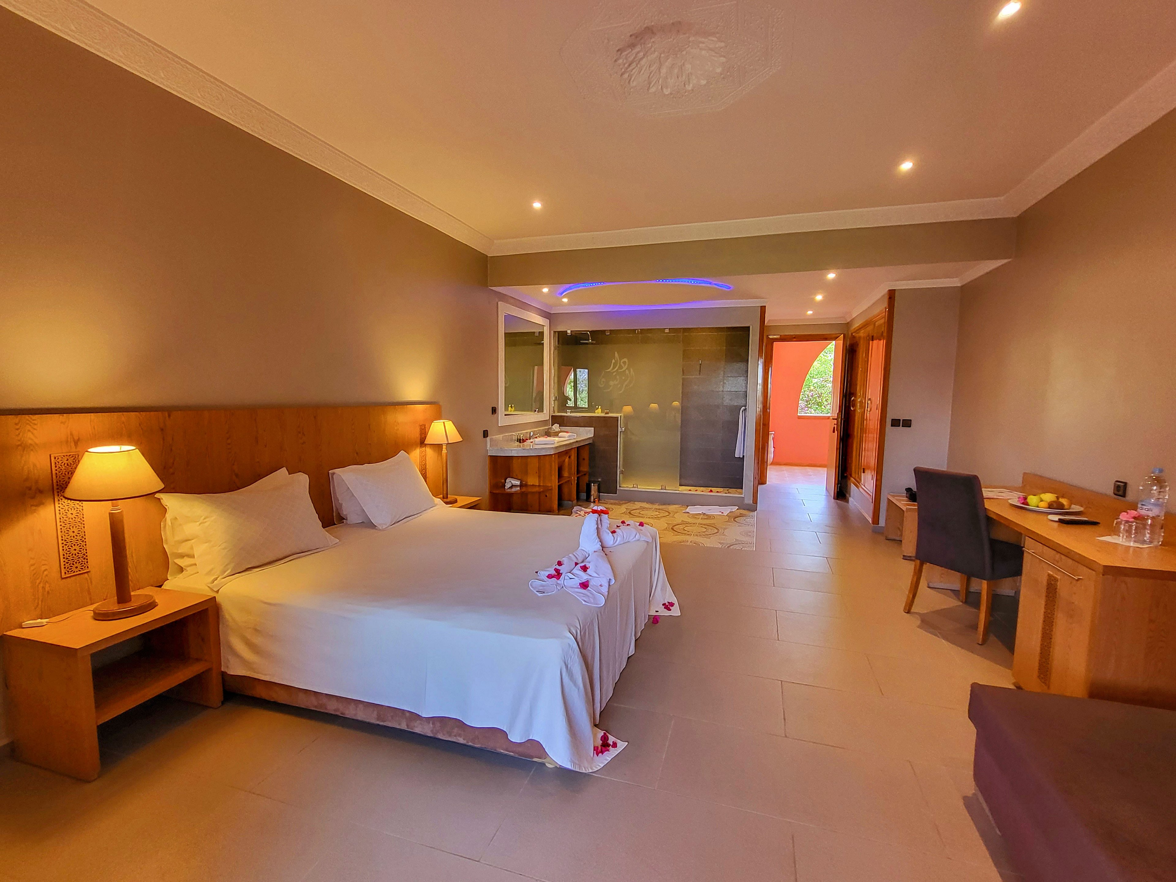 A warm, spacious hotel room features a neatly made double bed with bedside lamps and wooden furniture. Towels folded into decorative shapes rest on the bed, adding a welcoming touch. At the far end, a large glass-walled bathroom area is visible, with soft ceiling lighting. A desk with a chair and refreshments sits along the wall, while an open doorway reveals an exterior view with greenery and orange walls.