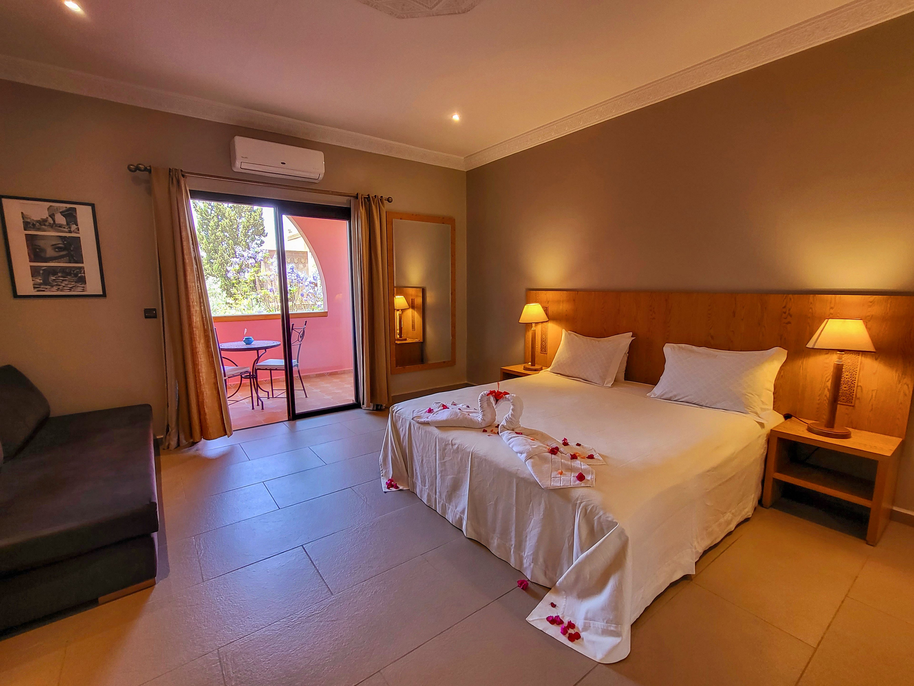 A warm, inviting hotel bedroom features a large double bed neatly made with white linens and decorative towel art. Soft lighting from bedside lamps creates a cozy atmosphere against neutral-colored walls. A sliding glass door opens to a balcony with outdoor chairs, allowing natural light to enter the room. A dark sofa, framed wall art, and simple wooden furniture complete the comfortable, modern interior.