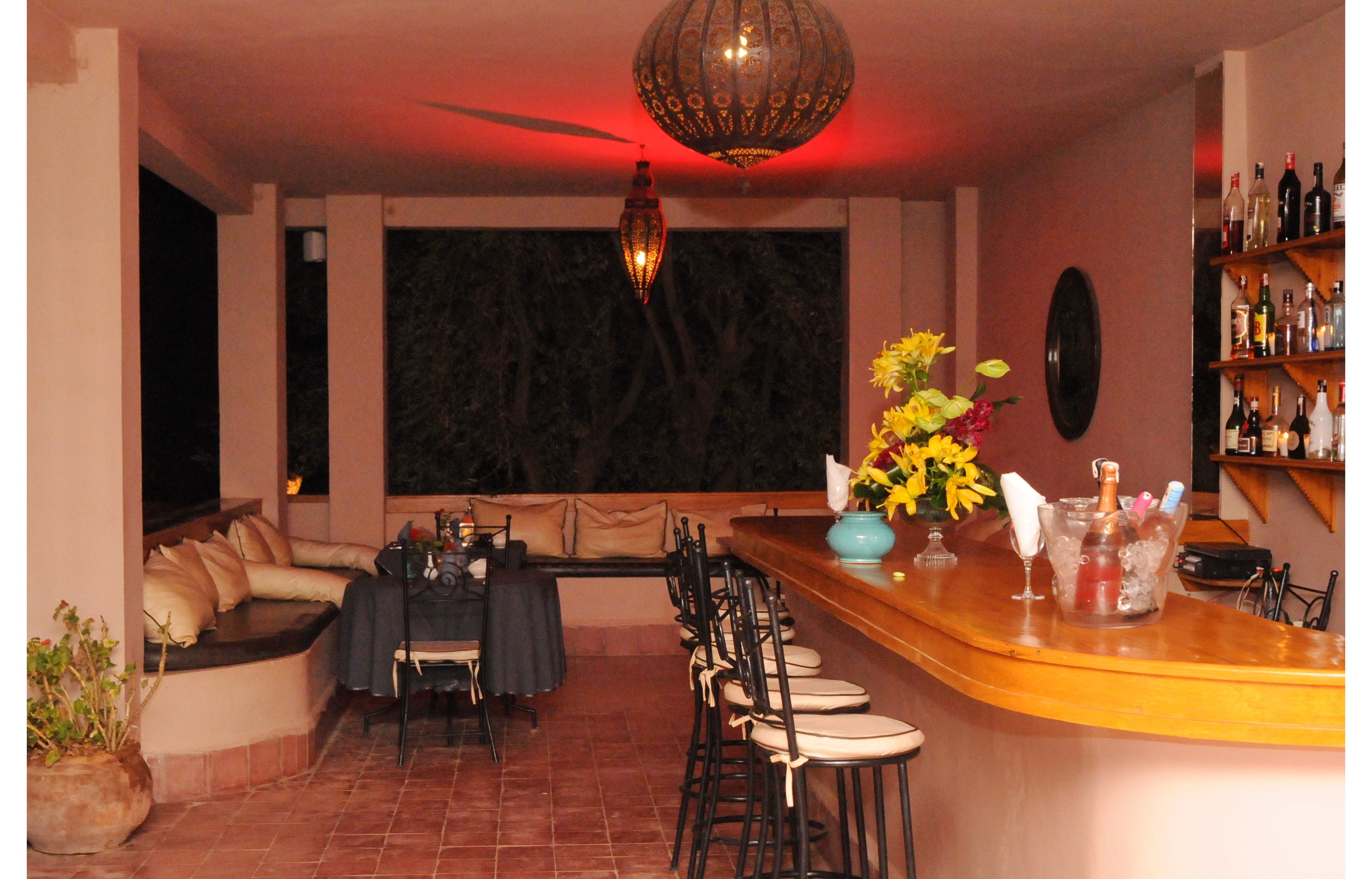 An intimate open-air bar is softly lit with warm ambient lighting during the evening. A curved wooden counter with barstools faces a wall-mounted shelf filled with assorted bottles. A small dining table is set with dark linens and folded napkins, while a cushioned bench seating area lines the side wall. Decorative pendant lamps and a vase of bright flowers add a welcoming touch to the relaxed atmosphere. The scene suggests a quiet, comfortable place for drinks and conversation.