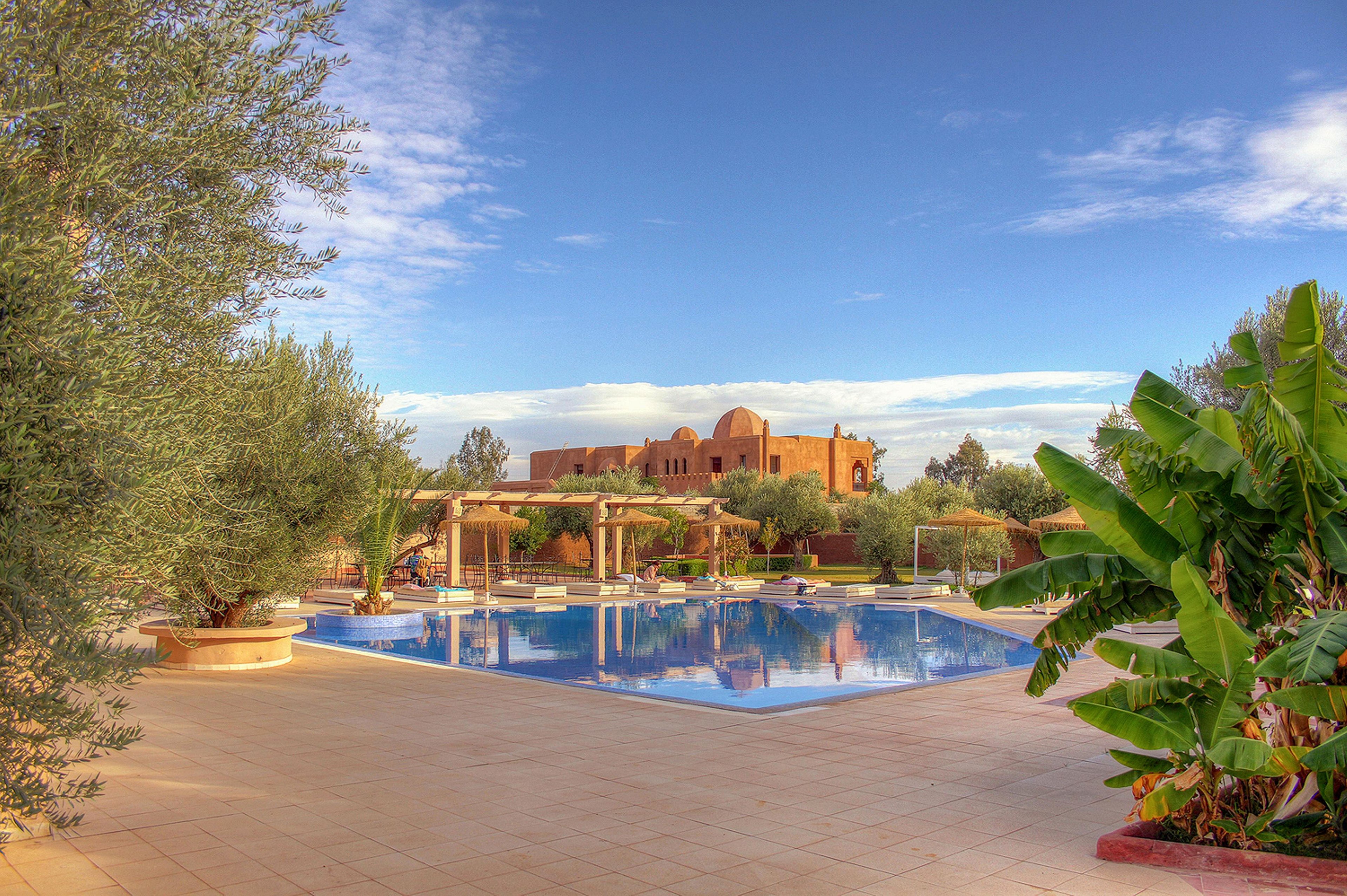 A large, tranquil swimming pool reflects the clear blue sky in front of a warm-toned, Mediterranean-style building. The scene is framed by lush greenery, including palm trees and other tropical plants, creating a relaxing resort atmosphere. Sun loungers and shaded pergolas surround the pool, suggesting a comfortable outdoor leisure area. The soft, golden light enhances the peaceful mood of this vacation setting.