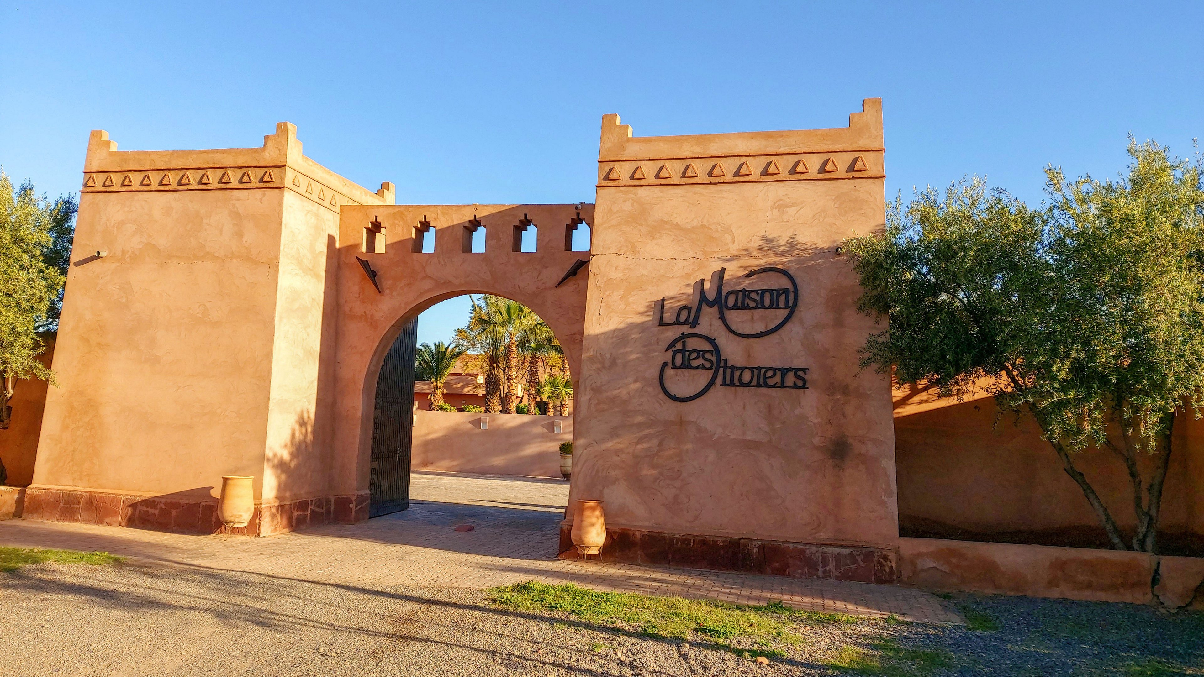 A large adobe-style gateway with an arched entrance stands under a clear blue sky. The structure features decorative crenellations along the top and warm earthy tones that catch the sunlight. On the right side of the gate, the text "La Maison des Oliviers" is clearly visible on the wall. Olive trees frame the entrance, and a gravel path leads toward the interior courtyard beyond the arch.
