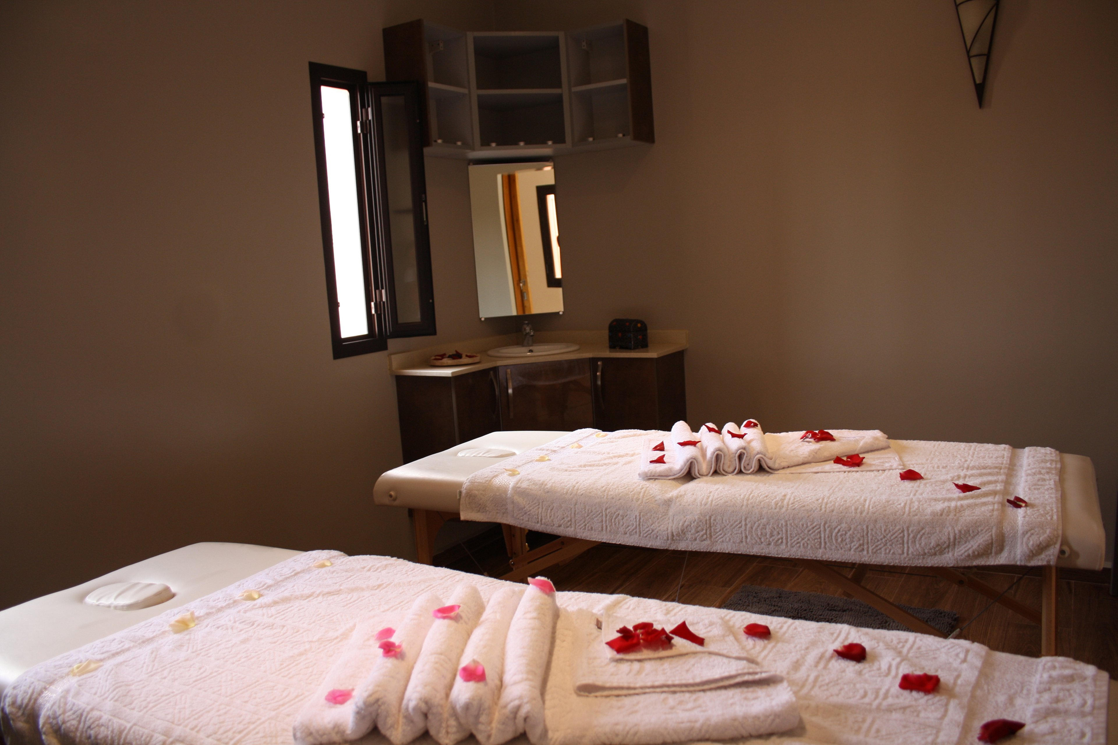 A softly lit spa treatment room features two massage tables neatly prepared with white towels. Rose petals are scattered across the linens, suggesting a romantic or couples treatment. An open window and a small vanity with a mirror appear in the background, adding natural light and depth to the space. The overall scene conveys relaxation, intimacy, and a serene wellness atmosphere.