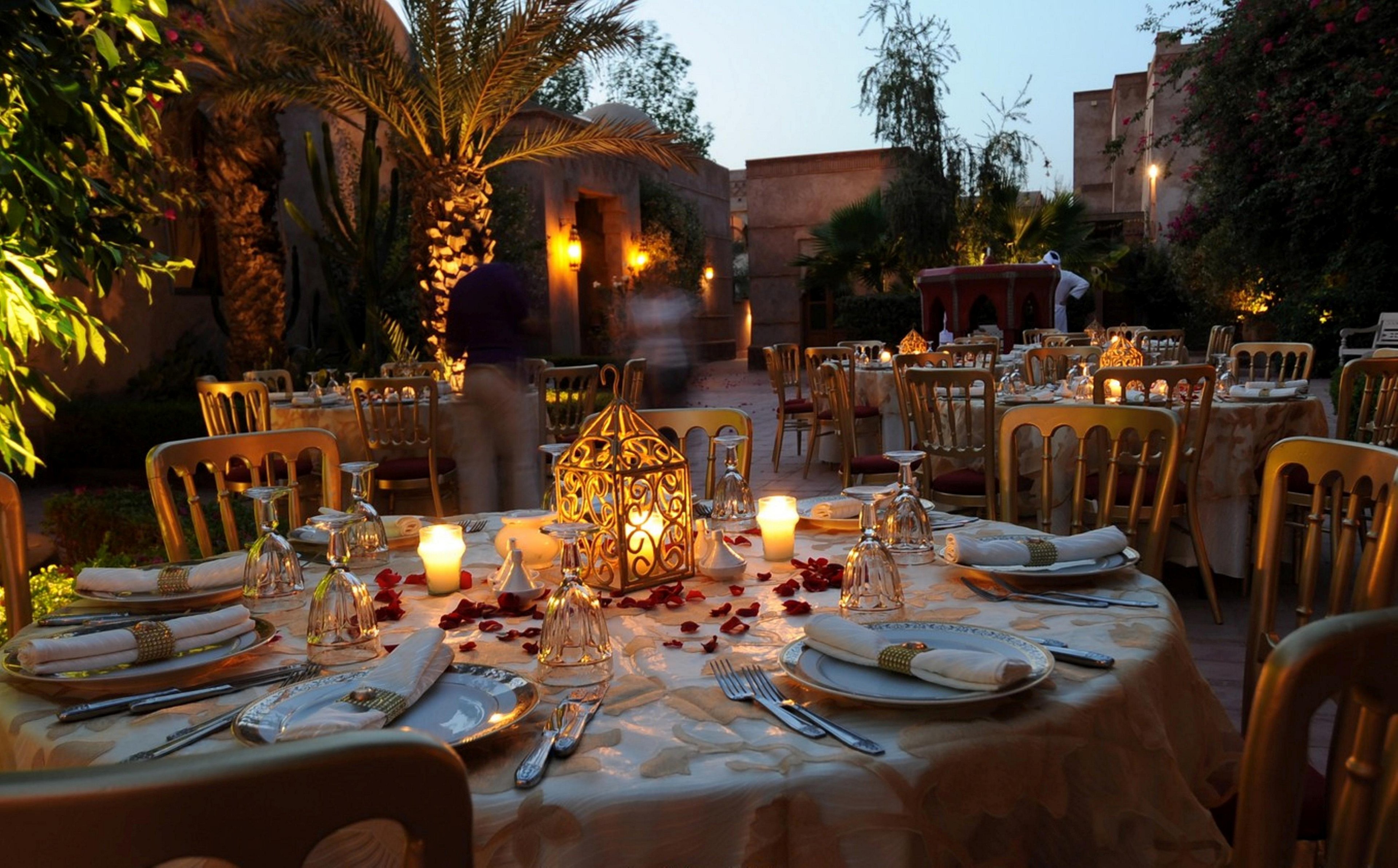 An elegantly set round table is arranged outdoors at dusk, featuring a decorative lantern centerpiece surrounded by candles and rose petals. Multiple tables with white linens and gold chairs fill the garden courtyard, creating an intimate dining atmosphere. Warm ambient lighting from wall sconces and lanterns illuminates palm trees and lush greenery in the background. Blurred figures suggest guests moving through the scene, enhancing the lively yet romantic mood.