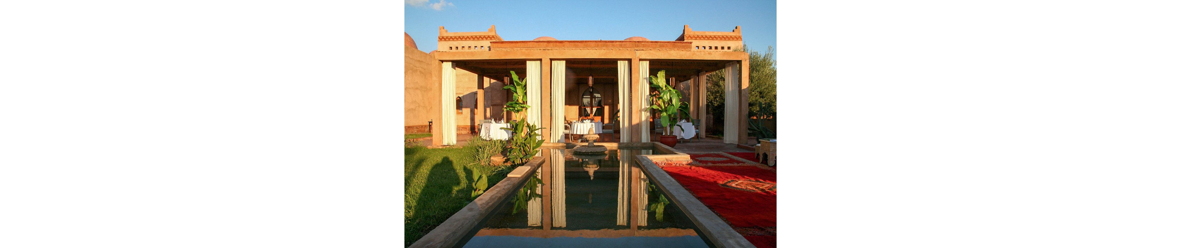 An open-air pavilion with draped columns stands at the end of a long reflecting pool. Dining tables with white tablecloths are set under the structure, suggesting an elegant outdoor restaurant or event space. Lush green plants and manicured grass contrast with warm terracotta walls and red carpets. The scene is bathed in golden sunlight, creating a tranquil and inviting atmosphere.