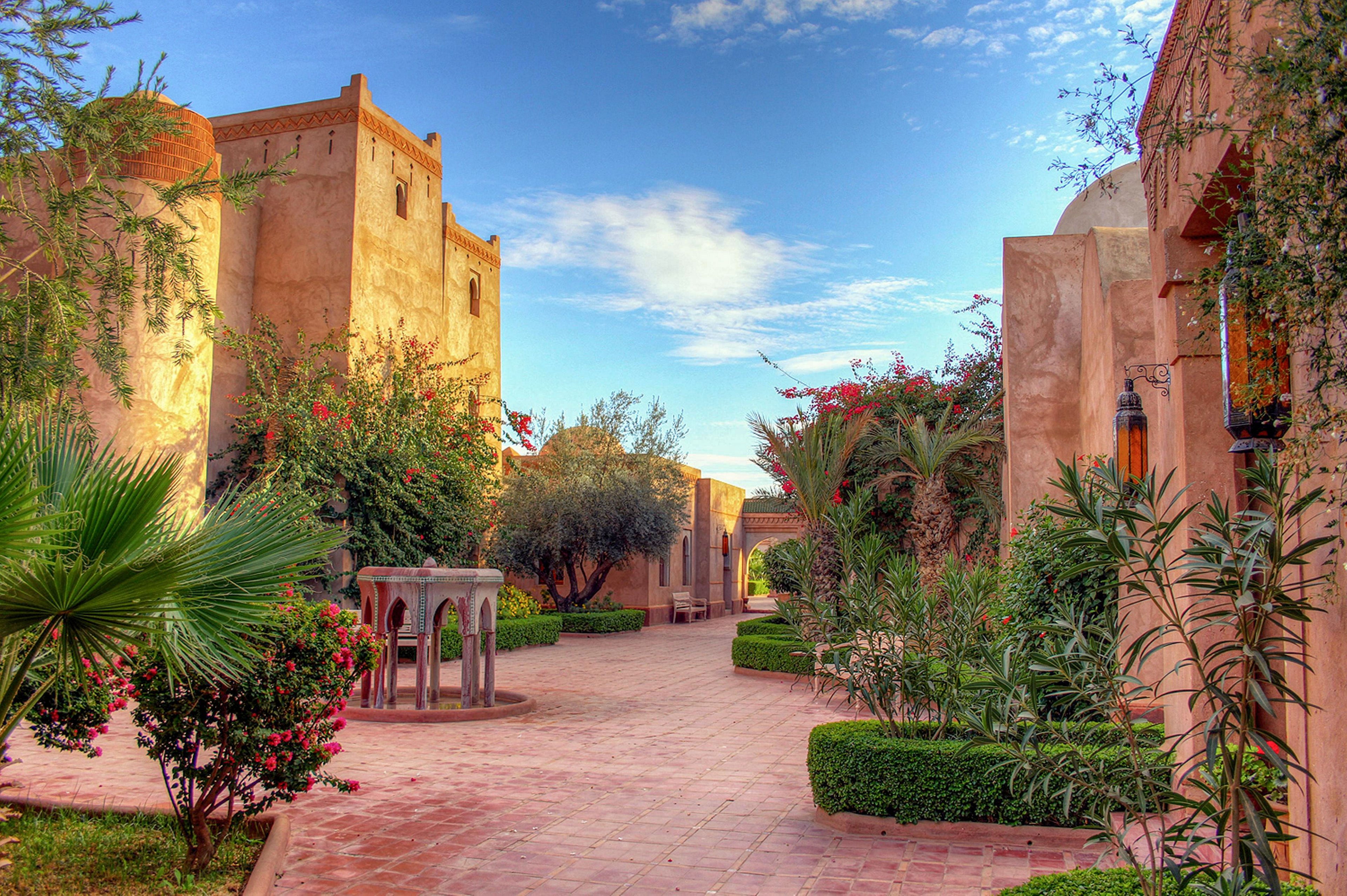 A quiet courtyard is lined with traditional adobe-style buildings under a bright blue sky with scattered clouds. The pathway is paved with warm-toned tiles and bordered by manicured hedges and lush green plants. Ornamental lanterns and a stone fountain add decorative detail to the serene outdoor space. The scene evokes a peaceful, inviting atmosphere in a warm, sunny climate.