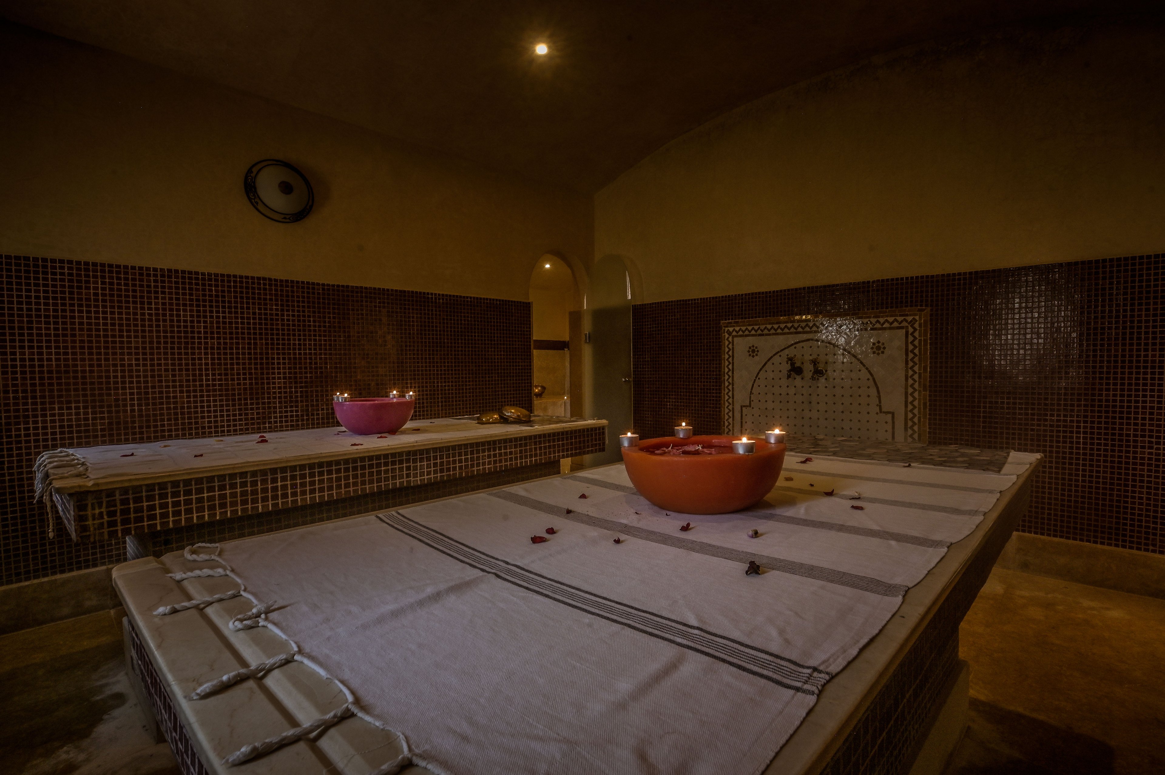 A tranquil spa treatment room features two stone massage tables covered with light-colored linens. Large bowls filled with water and rose petals sit at the center of each table, surrounded by small lit candles that create a warm glow. The walls are tiled in a rich, dark tone, with an arched doorway and decorative niche adding architectural interest. The dim lighting and scattered petals evoke a calm, relaxing atmosphere ideal for wellness and rejuvenation.