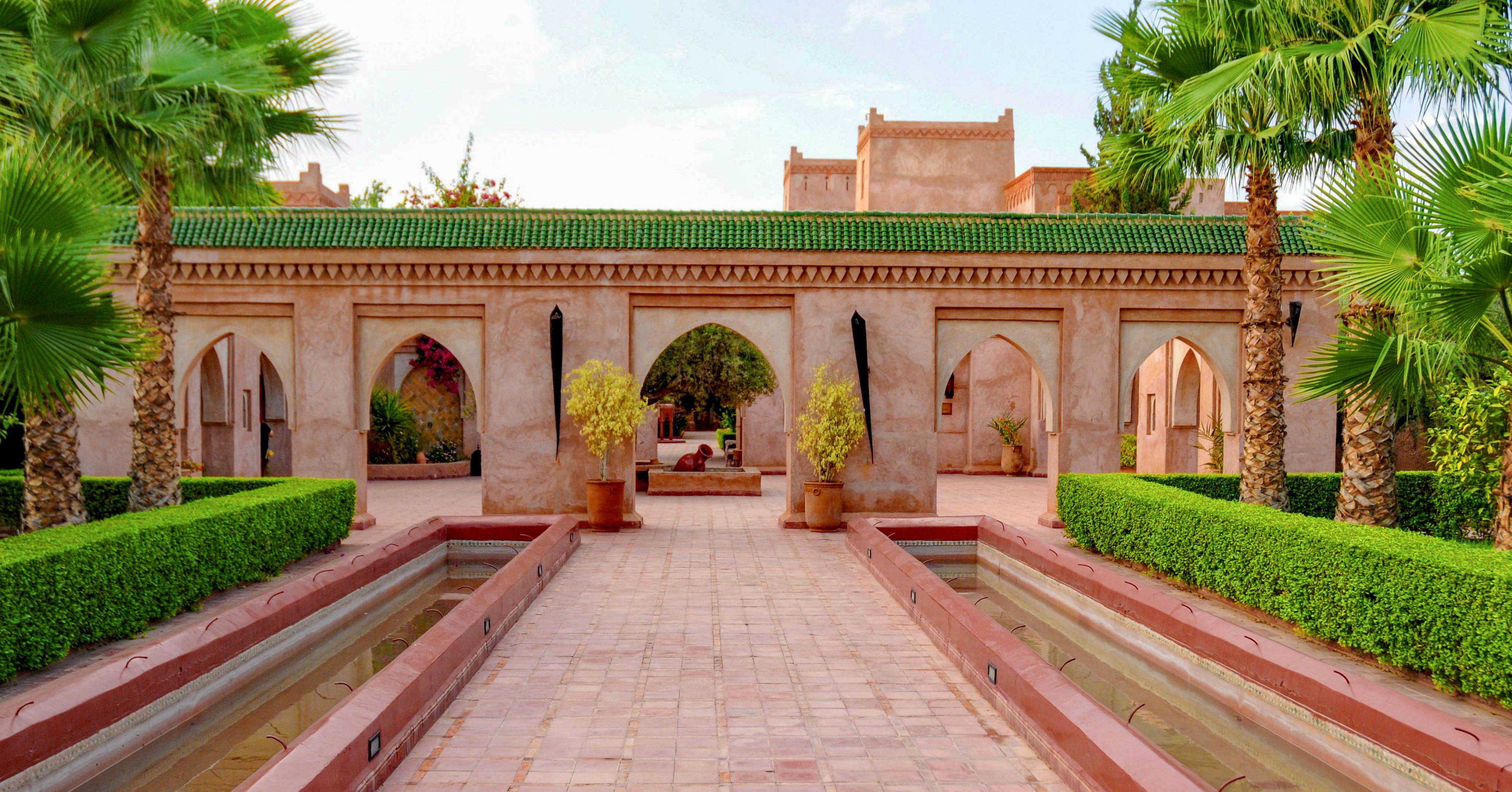 An elegant courtyard features a long tiled walkway flanked by narrow water channels. The central building showcases multiple arches and a green-tiled roof, evoking traditional architectural design. Manicured hedges and tall palm trees line the path, adding vibrant greenery to the warm stone tones. The scene appears calm and inviting, suggesting a tranquil garden or resort setting.