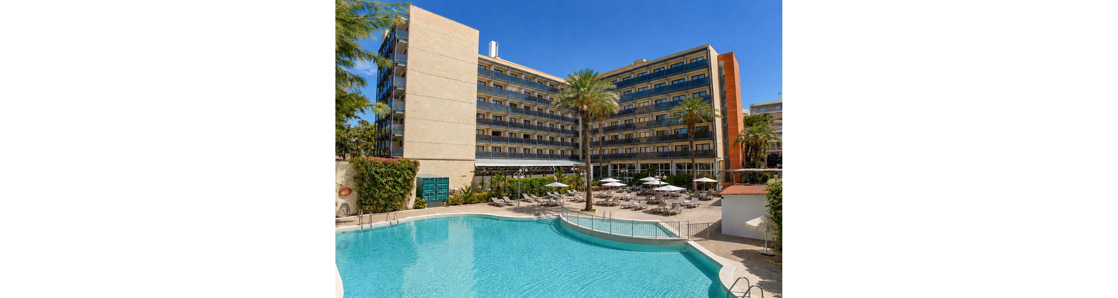 A multi-story modern hotel overlooks a large outdoor swimming pool under a clear blue sky. The pool area features sun loungers, umbrellas, and seating arranged neatly around the water. Tall palm trees and green hedges add a tropical touch to the otherwise urban architecture. The bright daylight and calm water create a relaxed vacation atmosphere.