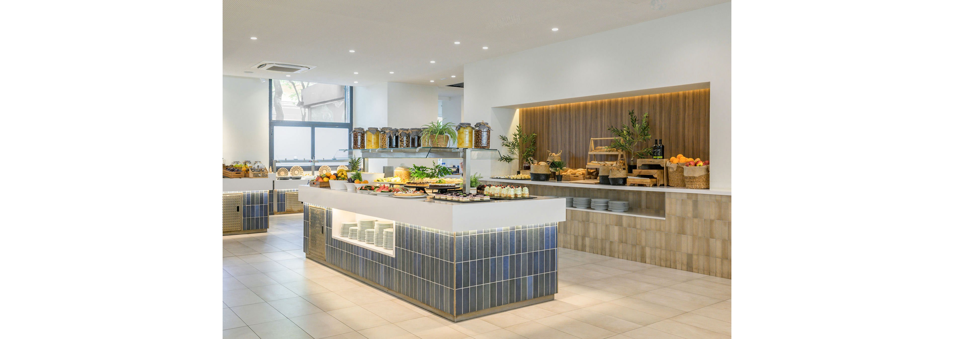A bright, modern buffet area showcases a wide selection of breakfast foods arranged on a central island counter. Glass jars of cereals and condiments line the back counter, while baskets of fresh fruit and bread sit on wooden stands. Neutral tiles, clean white surfaces, and integrated lighting create a contemporary, minimalist atmosphere. Large windows allow natural light to fill the space, highlighting the organized and inviting food display.