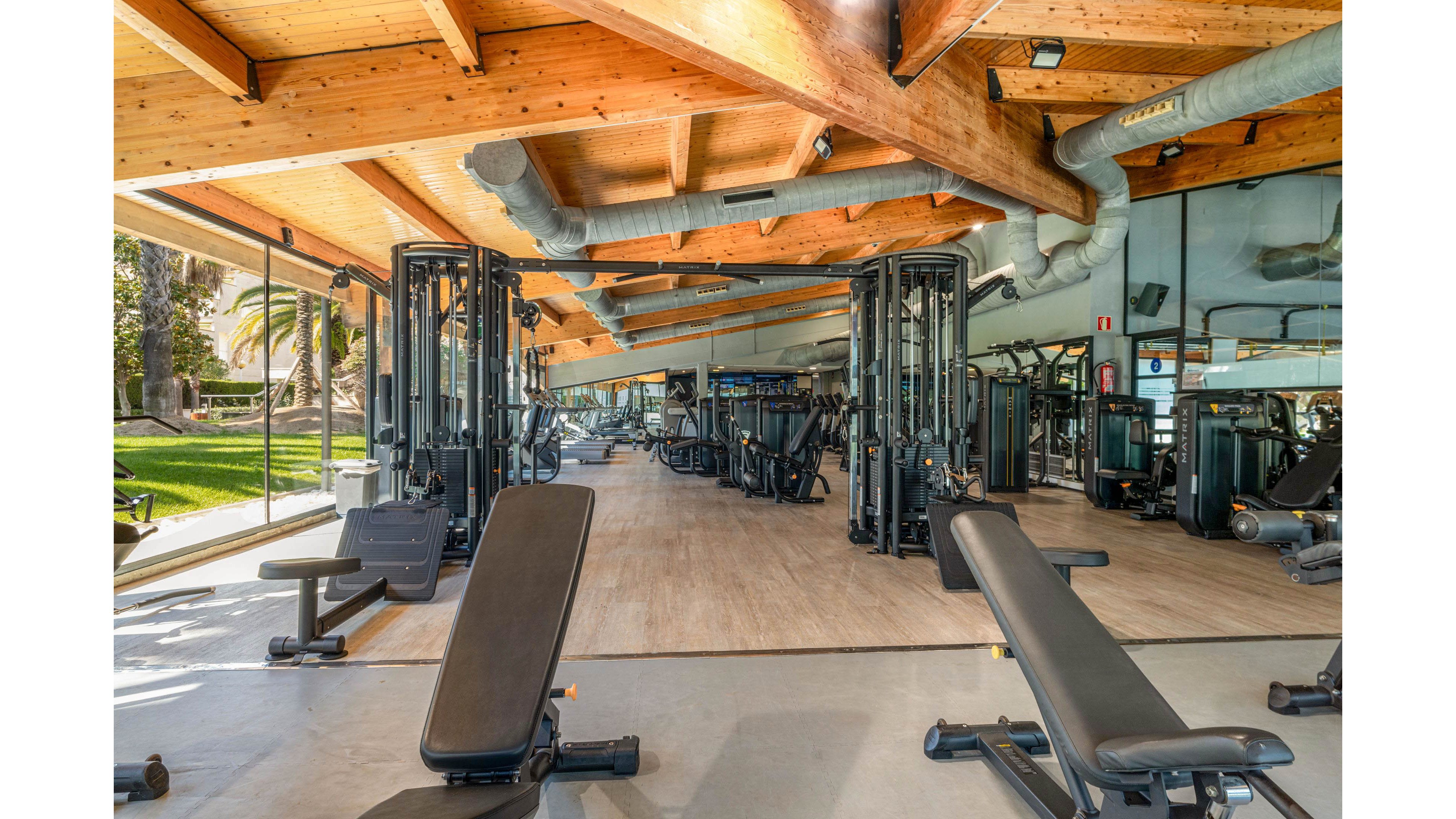 Spacious fitness center featuring multiple weight benches and cable machines under a wooden ceiling. Large windows and open sides reveal a green outdoor area with palm trees, creating an airy, resort-like atmosphere. The gym floor is clean and organized, with equipment arranged in rows for strength training. Natural light and reflective surfaces enhance the contemporary, professional workout environment.