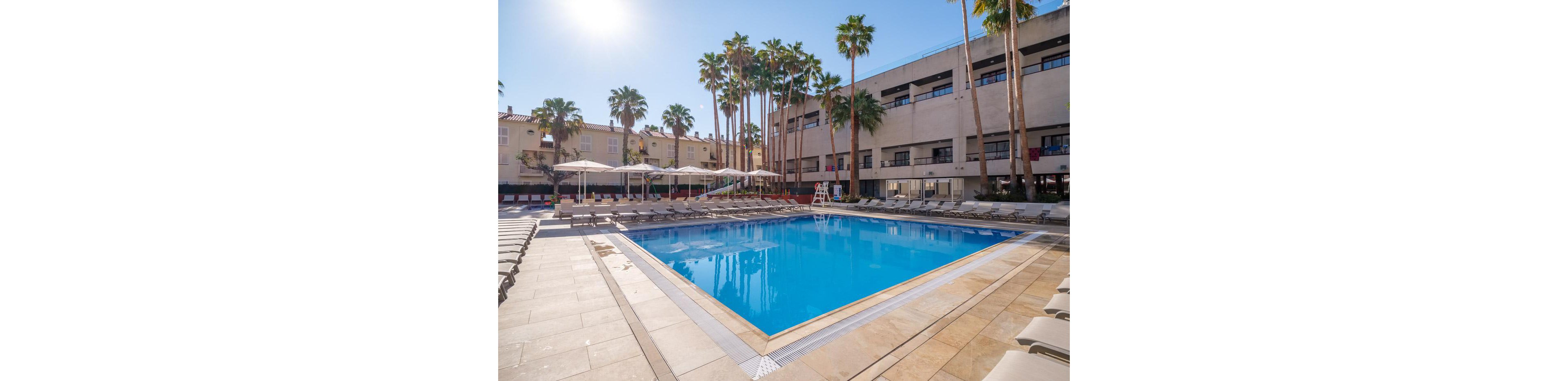 A large outdoor swimming pool is surrounded by lounge chairs and umbrellas in a resort courtyard. Tall palm trees rise above the pool area, adding a tropical atmosphere to the scene. A lifeguard chair stands near the pool, and multi-story buildings frame the background. The bright blue water and clear sky create a warm, inviting vacation setting.
