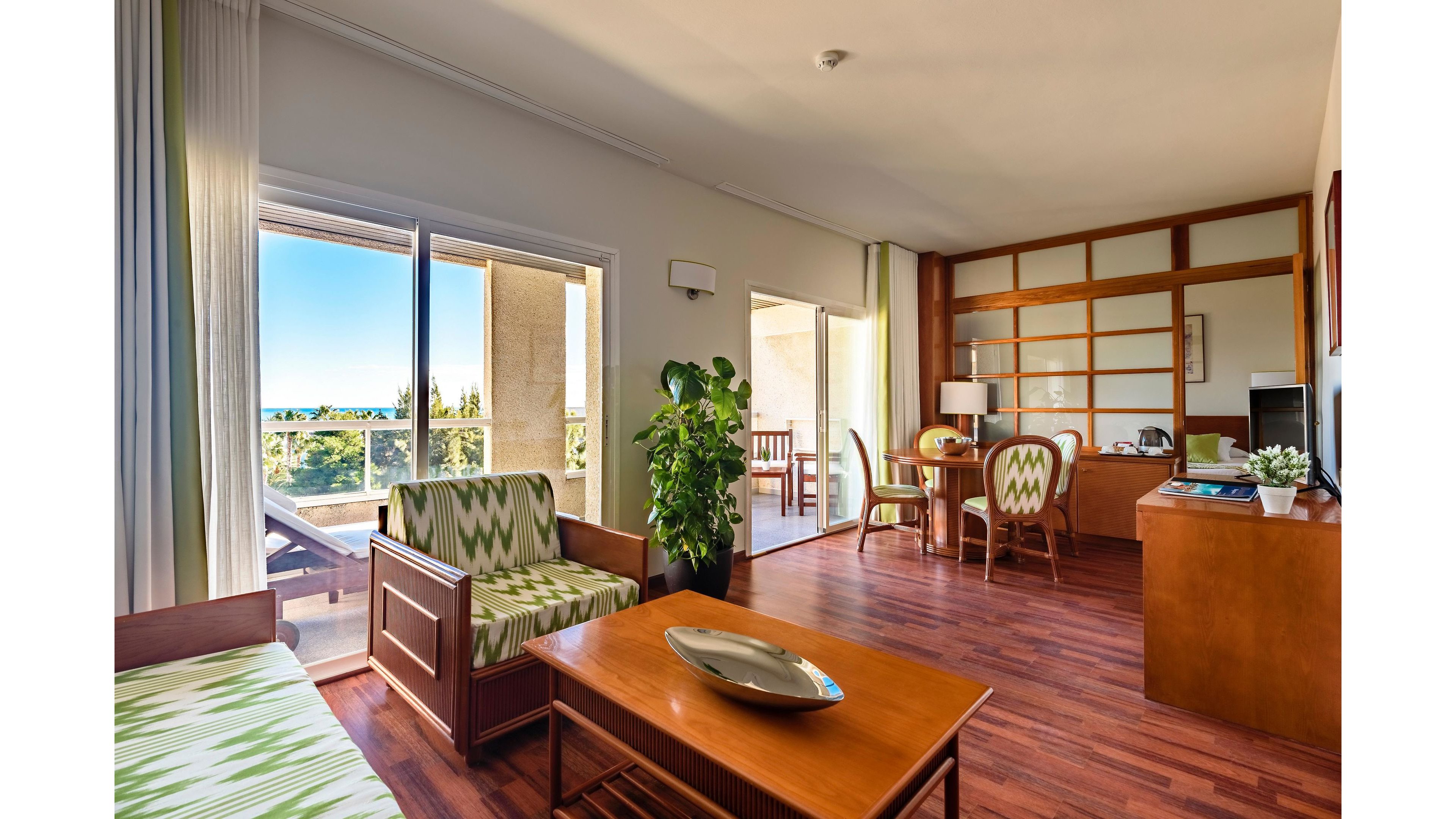 Spacious hotel suite featuring a living area with wooden furniture and patterned green cushions. Large sliding glass doors open to a balcony with lounge chairs and outdoor seating, overlooking a sunny landscape. The room includes a small dining table, desk area, and indoor plant, all set on warm wood flooring. Natural light and neutral walls create an airy, inviting atmosphere ideal for vacation stays.