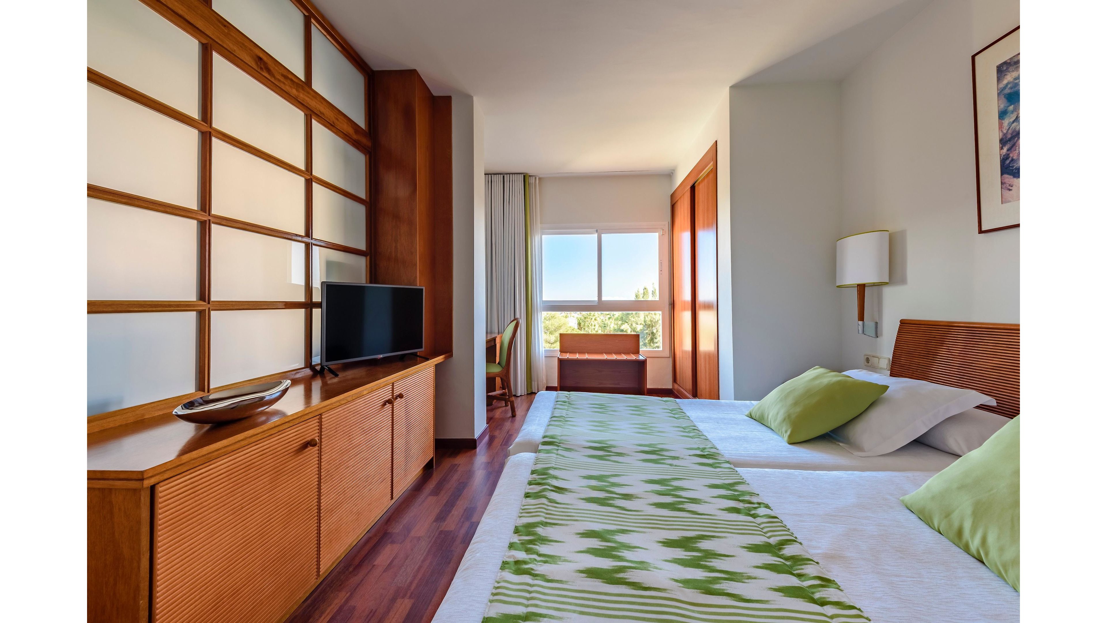 Contemporary hotel room featuring two neatly made twin beds with green patterned runners and matching pillows. The space includes warm wooden furniture, a flat-screen television on a low cabinet, and a desk near the window. Large windows let in natural daylight and offer a view of trees and distant buildings. Overall, the room feels clean, inviting, and designed for comfortable vacation or business stays.