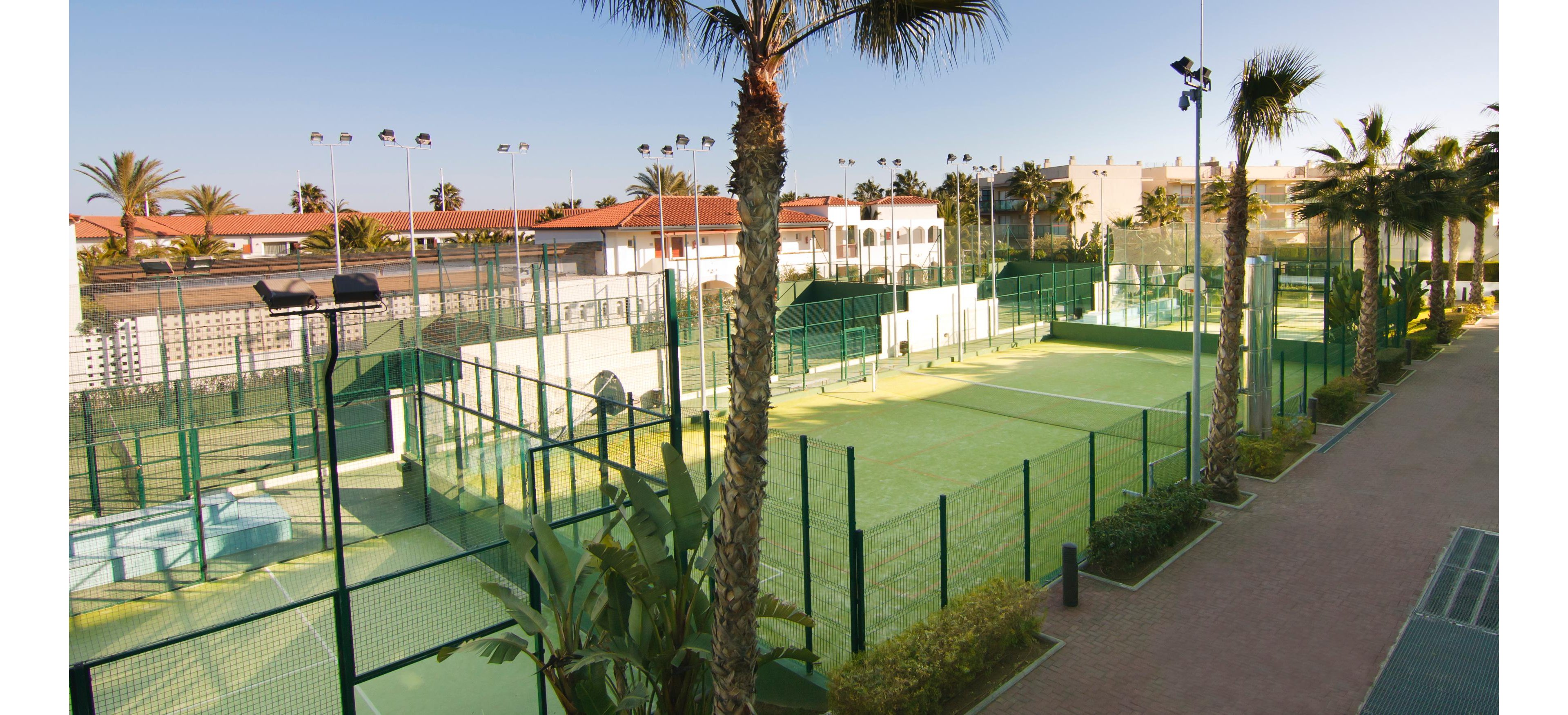 Image of multiple outdoor padel tennis courts surrounded by high green fences. The courts are located in a resort area with palm trees and low-rise buildings in the background. Bright sunlight and clear skies create a warm, vacation atmosphere. A paved walkway runs alongside the courts, adding to the organized sports facility setting.