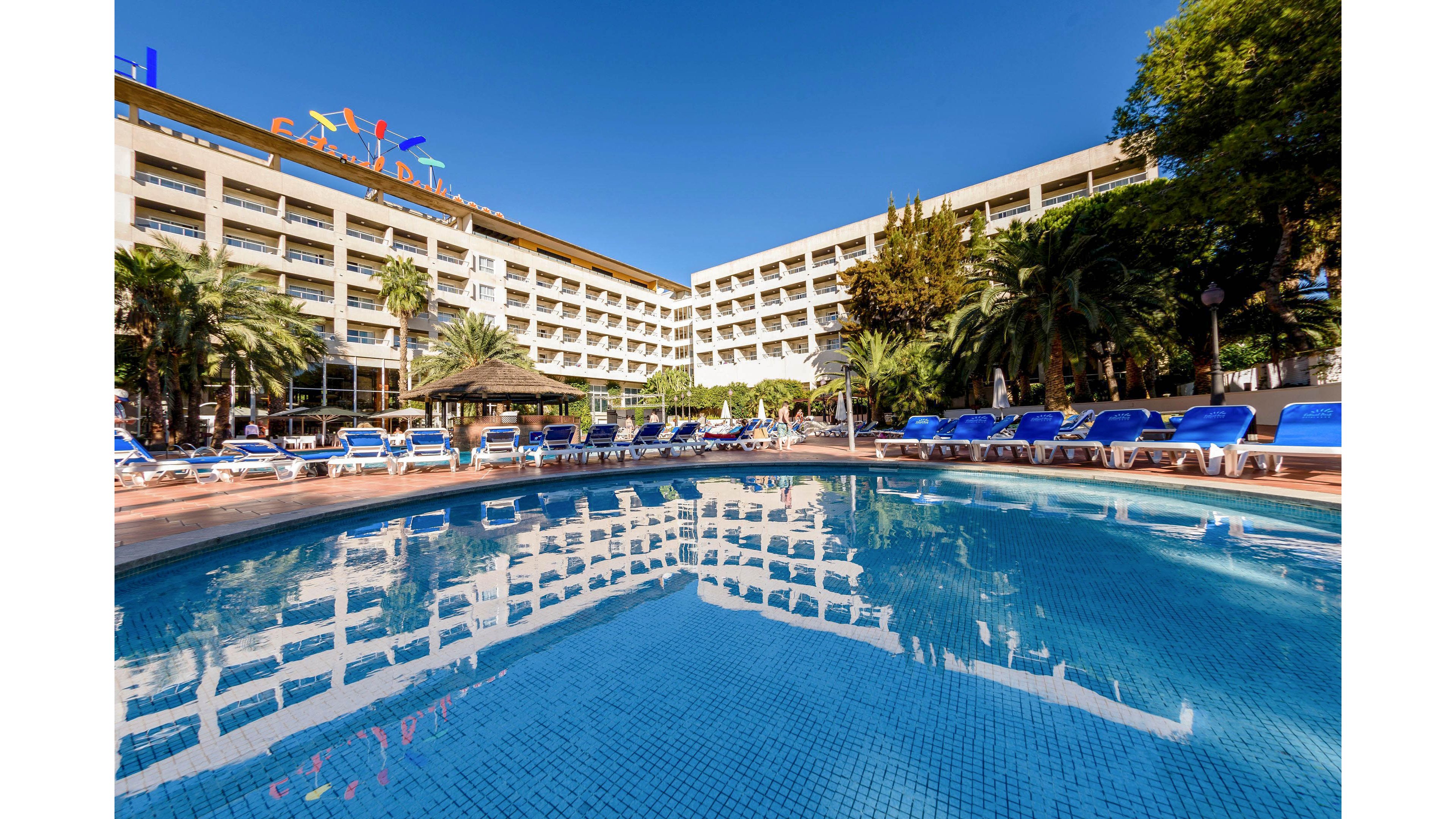 A large resort hotel rises behind a clear blue outdoor swimming pool under a bright, cloudless sky. Rows of blue sun loungers and umbrellas surround the pool, suggesting a relaxing vacation setting. Palm trees and lush greenery frame the scene, adding a tropical atmosphere. The hotel facade features multiple balconies and colorful signage at the rooftop, emphasizing a lively holiday destination.