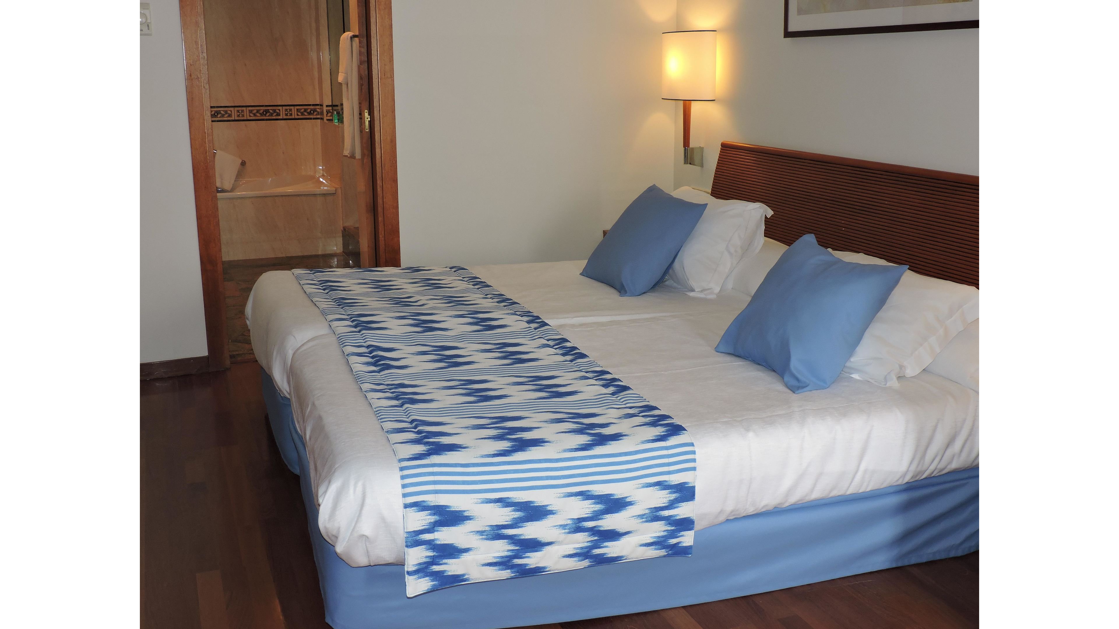 A neatly made double bed with white linens and blue pillows is the main focus of this hotel room. A patterned blue and white runner decorates the foot of the bed, complementing the blue bed skirt. Warm wall lighting and wooden flooring create a comfortable, inviting atmosphere. An open doorway reveals a tiled bathroom area in the background.