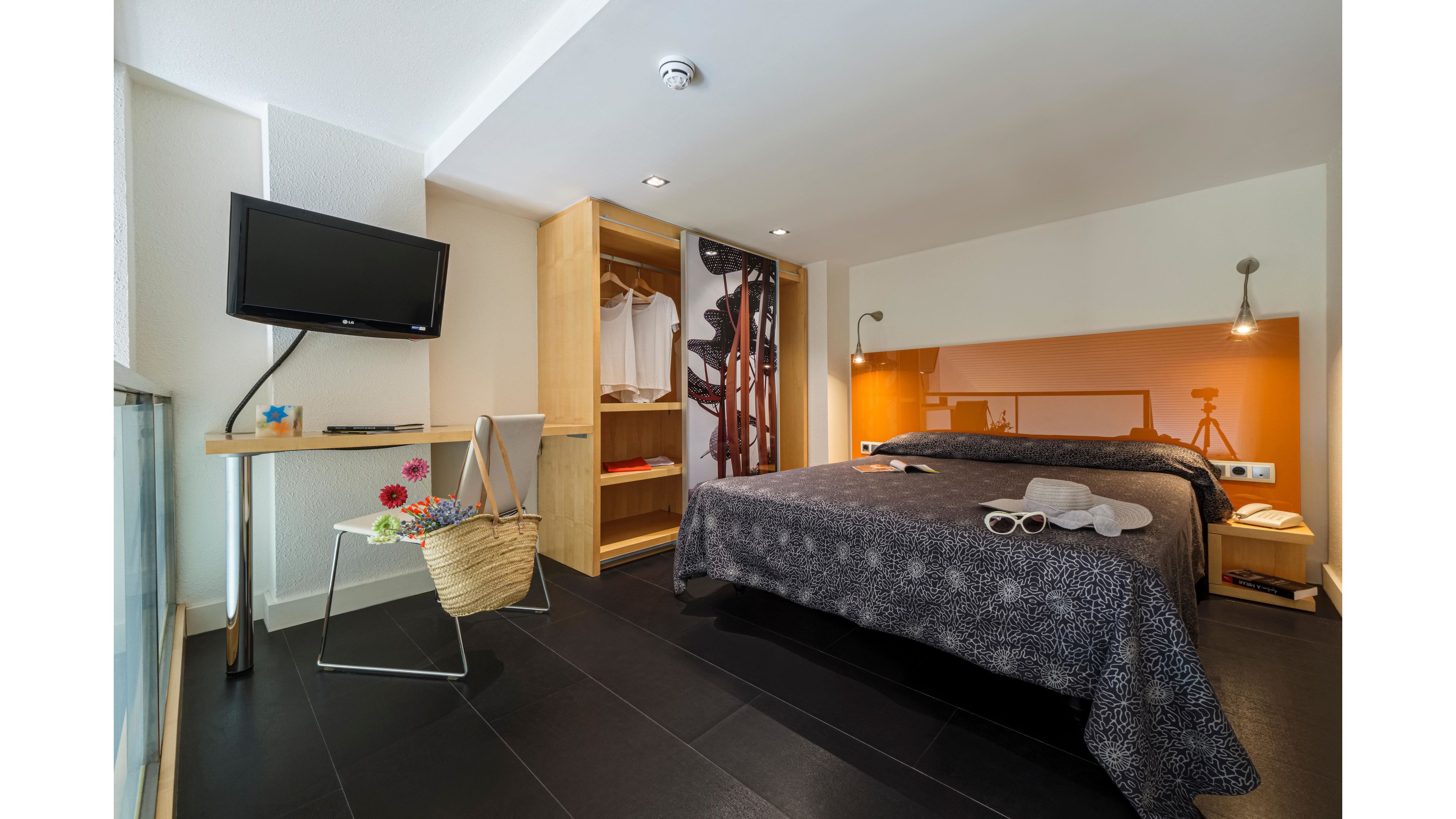 Contemporary hotel room features a double bed with a dark patterned bedspread and bedside lamps. An open wooden wardrobe displays hanging clothes and shelves, adding to the organized feel of the space. A flat-screen television is mounted on the wall opposite the bed, and a small side table holds flowers and a woven bag. Warm accent lighting and orange wall paneling create a cozy, inviting atmosphere.