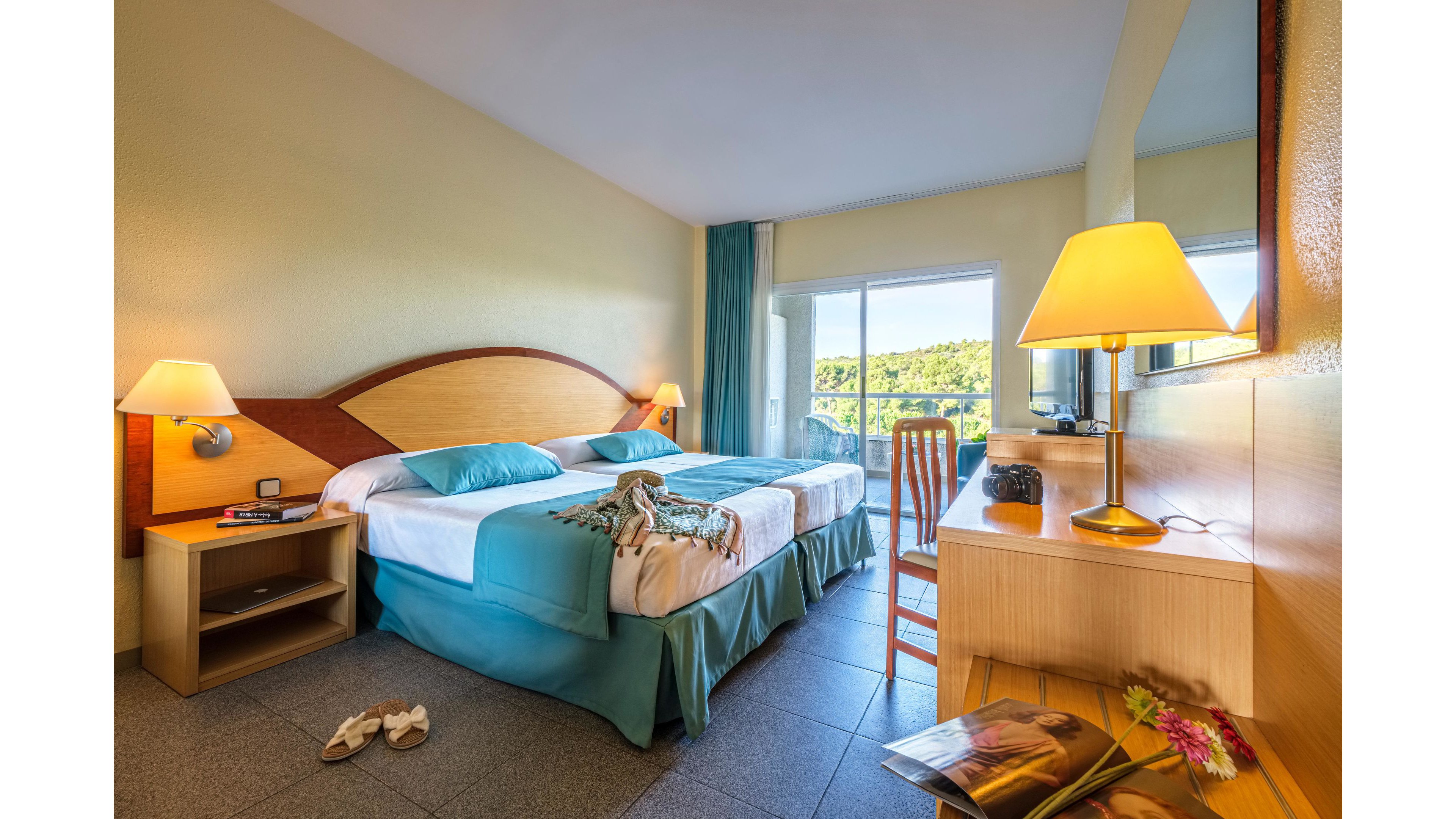 A sunlit hotel room features two neatly made twin beds with teal bedding and decorative pillows. A wooden desk with a lamp, chair, and television sits opposite the beds, adding functionality to the space. The room opens to a balcony through large glass doors, revealing greenery and a scenic outdoor view. Personal items like sandals, a scarf on the bed, and a magazine with flowers on the desk create a relaxed vacation atmosphere.