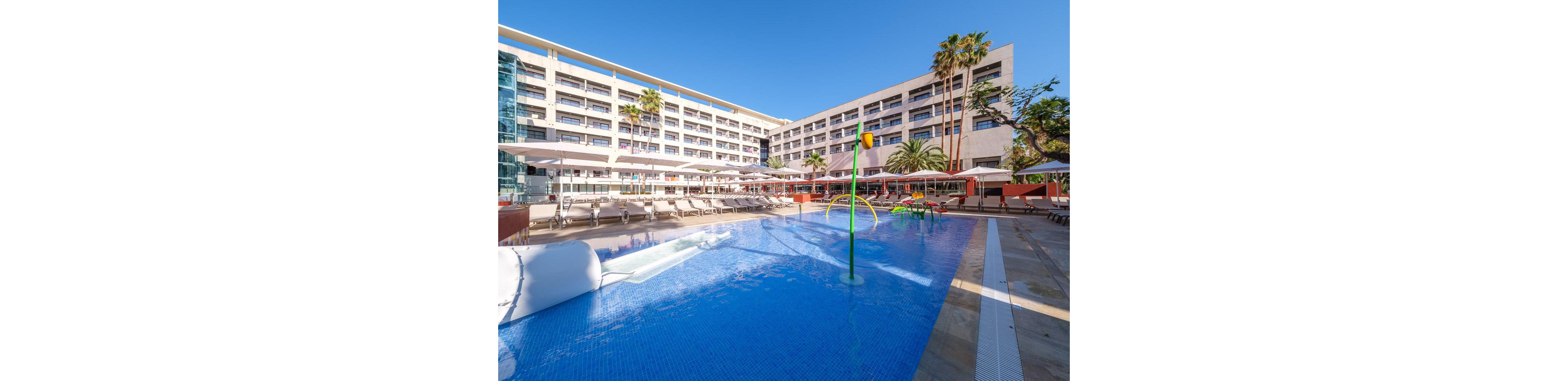 A large resort hotel features an expansive outdoor swimming pool in the foreground. Sun loungers and umbrellas line the poolside terrace, creating a relaxed vacation atmosphere. Colorful water play structures stand in the shallow pool, while tall palm trees add a tropical touch. The multi-story hotel building with balconies rises behind the pool under a clear blue sky.