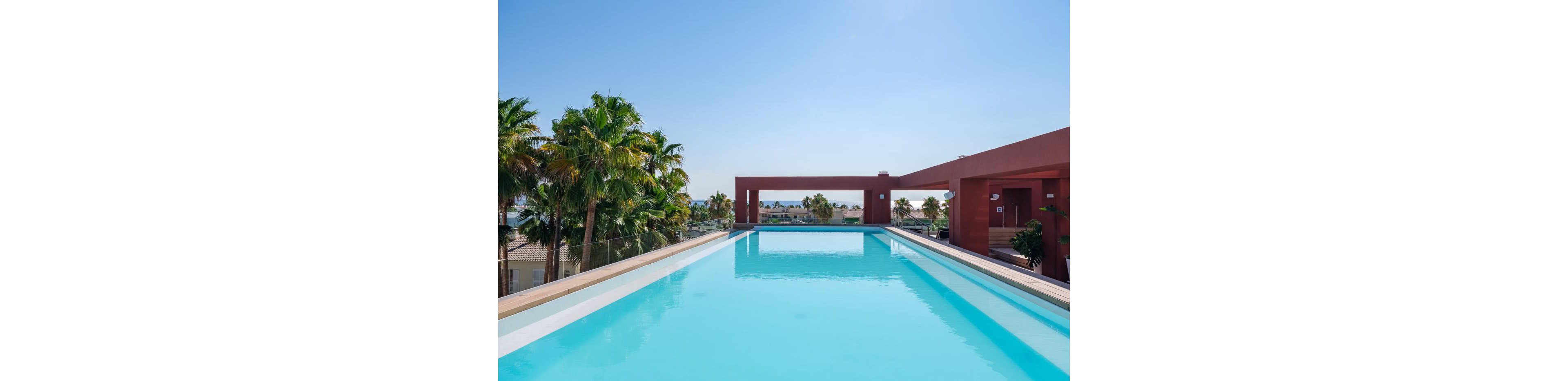 A long, modern rooftop swimming pool stretches toward an open rectangular frame overlooking a coastal town. Palm trees line the left side, adding a tropical feel to the contemporary architecture. Clear blue water reflects the bright sky, creating a serene and inviting atmosphere. In the distance, buildings and the sea are visible under sunny weather.