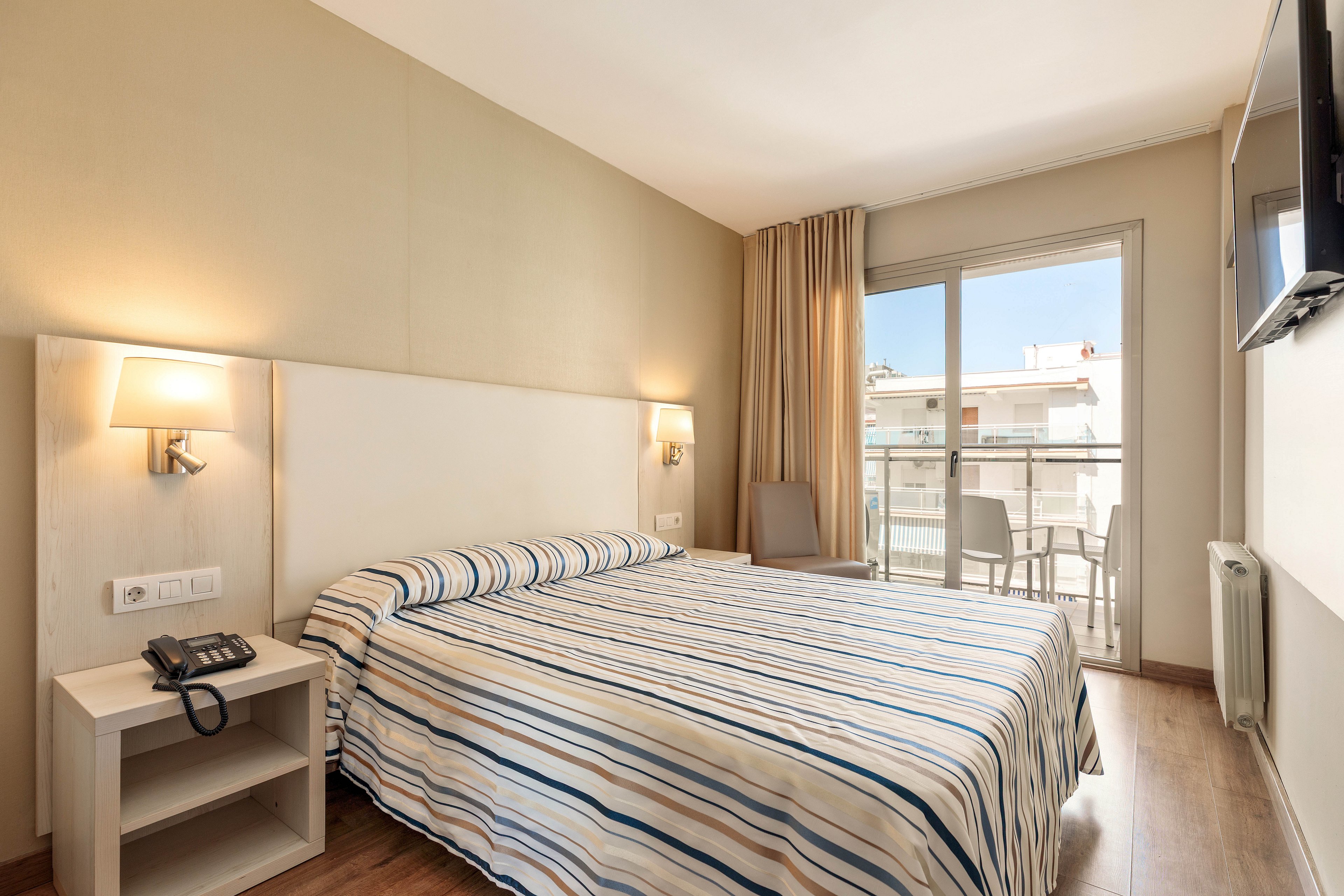 A neatly made double bed with a striped bedspread is the focal point of this bright hotel room. The space features light-colored walls, wooden flooring, and a bedside table with a telephone and wall-mounted lamp. Large sliding glass doors open onto a small balcony with outdoor chairs, allowing natural light to fill the room. A flat-screen TV is mounted on the wall opposite the bed, creating a comfortable and modern guest space.
