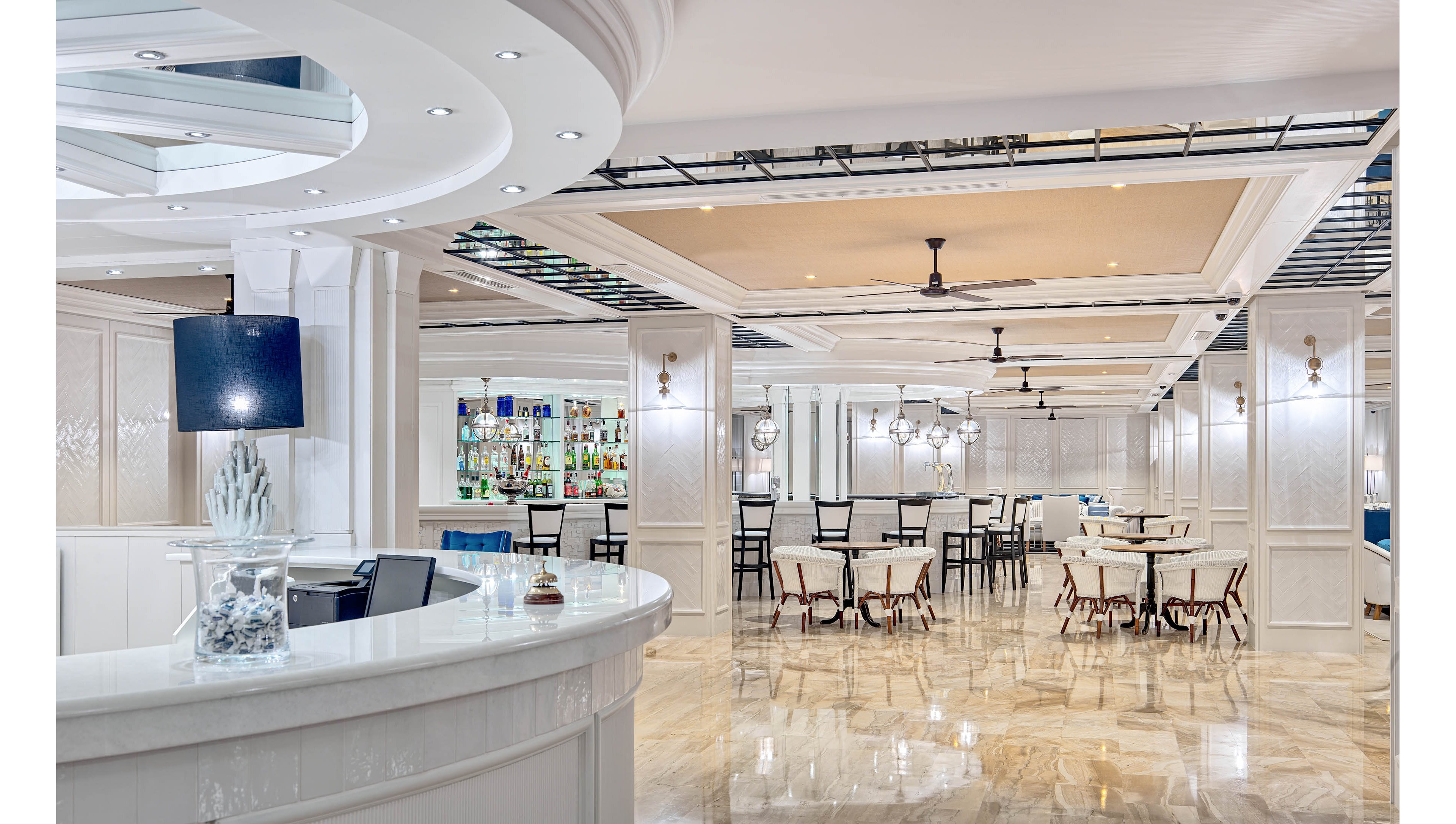 A spacious, contemporary hotel bar and dining lounge features glossy marble floors and white paneled walls. A curved reception or bar counter stands in the foreground, while a fully stocked bar with illuminated shelves is visible in the background. Multiple dining tables and chairs are neatly arranged under recessed ceiling panels with ceiling fans and modern light fixtures. The overall palette is light and neutral, creating an elegant and airy atmosphere.