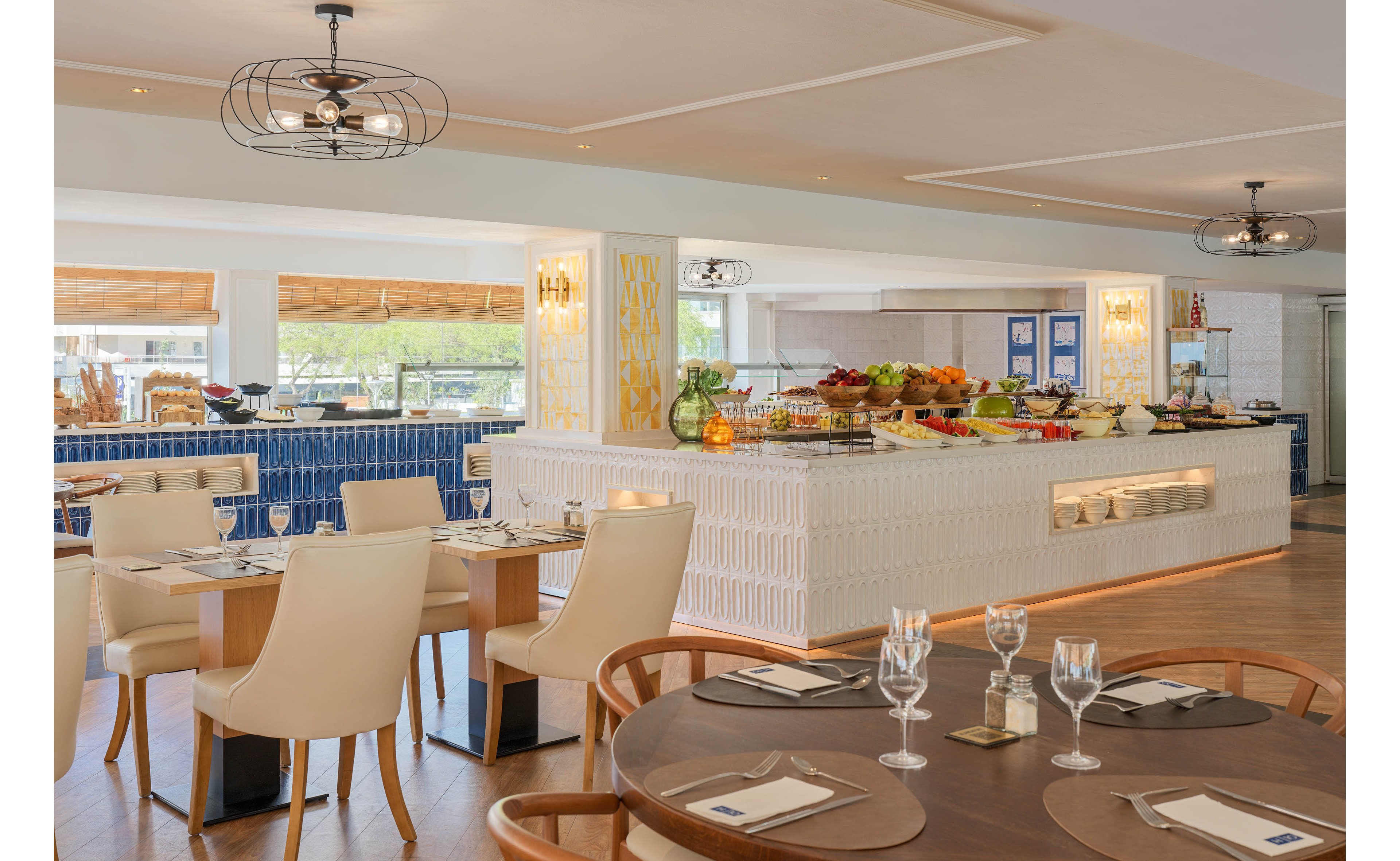 A contemporary hotel restaurant features a central buffet island filled with fresh fruits and dishes. Wooden tables with neatly arranged place settings and wine glasses surround the buffet area. Large windows bring in natural light and offer a view of greenery outside. Warm neutral tones, blue accents, and modern ceiling lights create an inviting dining atmosphere.