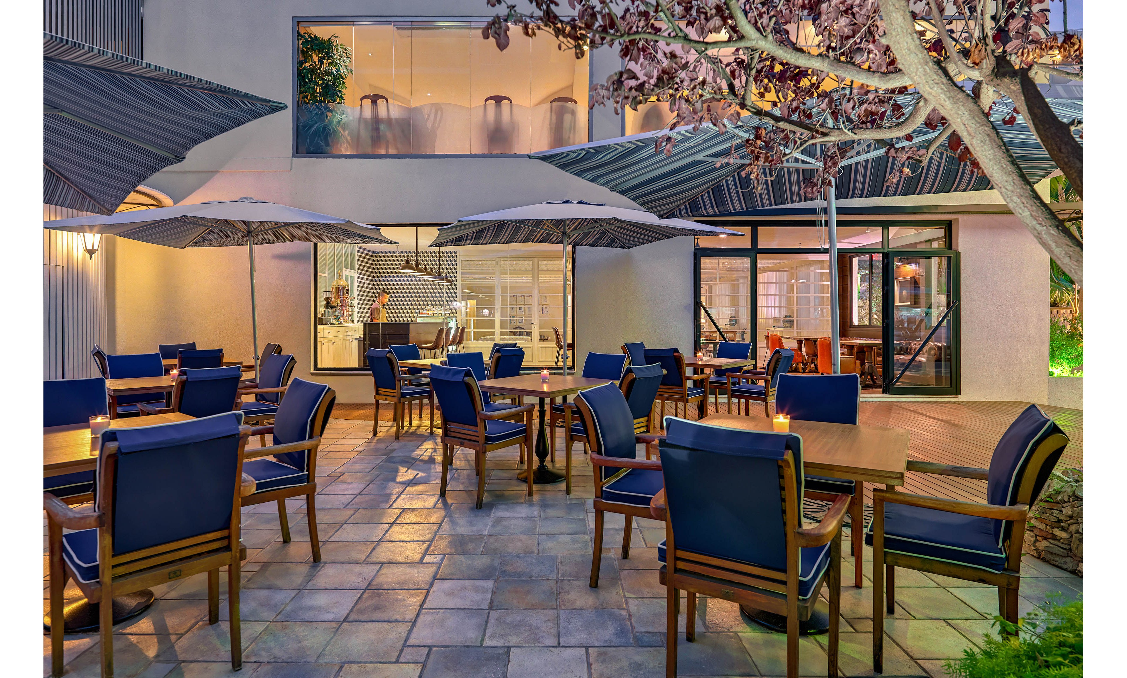 A contemporary outdoor dining patio features wooden tables and blue cushioned chairs neatly arranged under large umbrellas. The setting appears to be part of a restaurant or hotel, with large windows revealing a bright interior space. Soft evening lighting and clean tiled flooring create a welcoming atmosphere for guests. Trees and greenery frame the patio, adding a touch of nature to the modern design.
