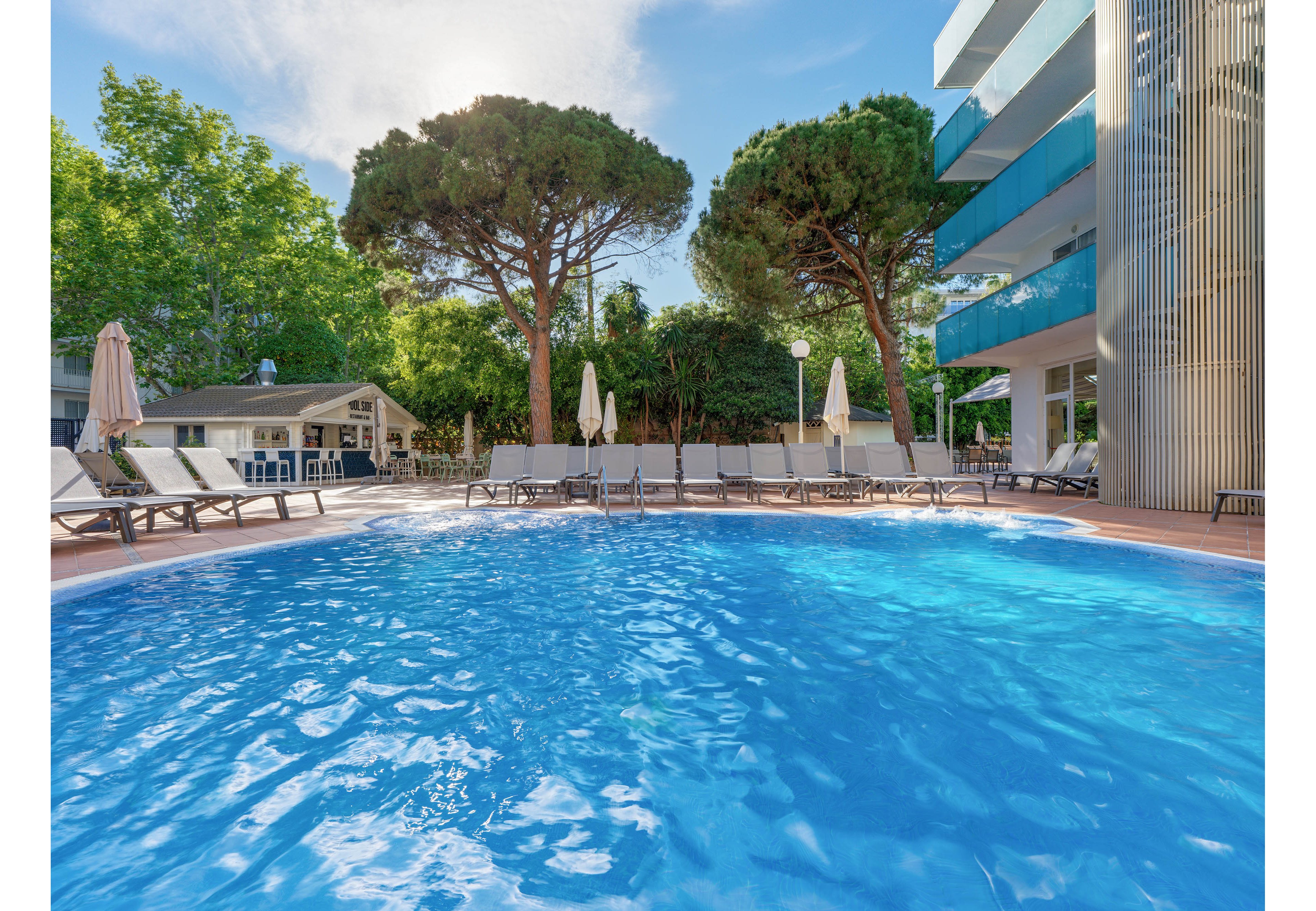 A bright blue swimming pool curves in the foreground of a modern hotel courtyard. Numerous empty lounge chairs and umbrellas line the poolside, suggesting a relaxing resort setting. Tall trees and lush greenery surround the area, adding shade and a natural backdrop. A contemporary multi-story building with balconies stands to the right, enhancing the vacation atmosphere under a clear sky.