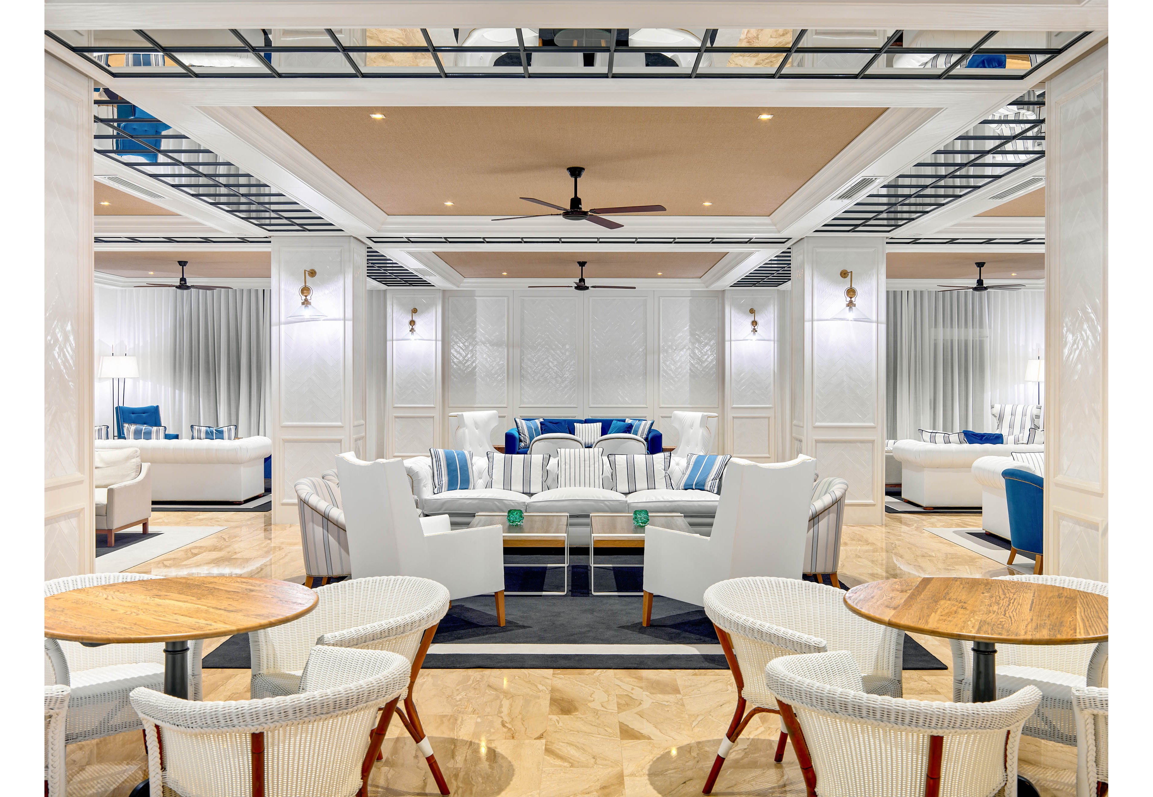 A bright, contemporary hotel lounge features multiple seating areas with white armchairs and round wooden tables. The space is designed with a neutral palette accented by blue cushions and decor, creating a coastal-inspired atmosphere. Ceiling fans, recessed lighting, and reflective ceiling panels add to the modern, airy feel. The polished floor and clean architectural lines emphasize sophistication and comfort for guests.
