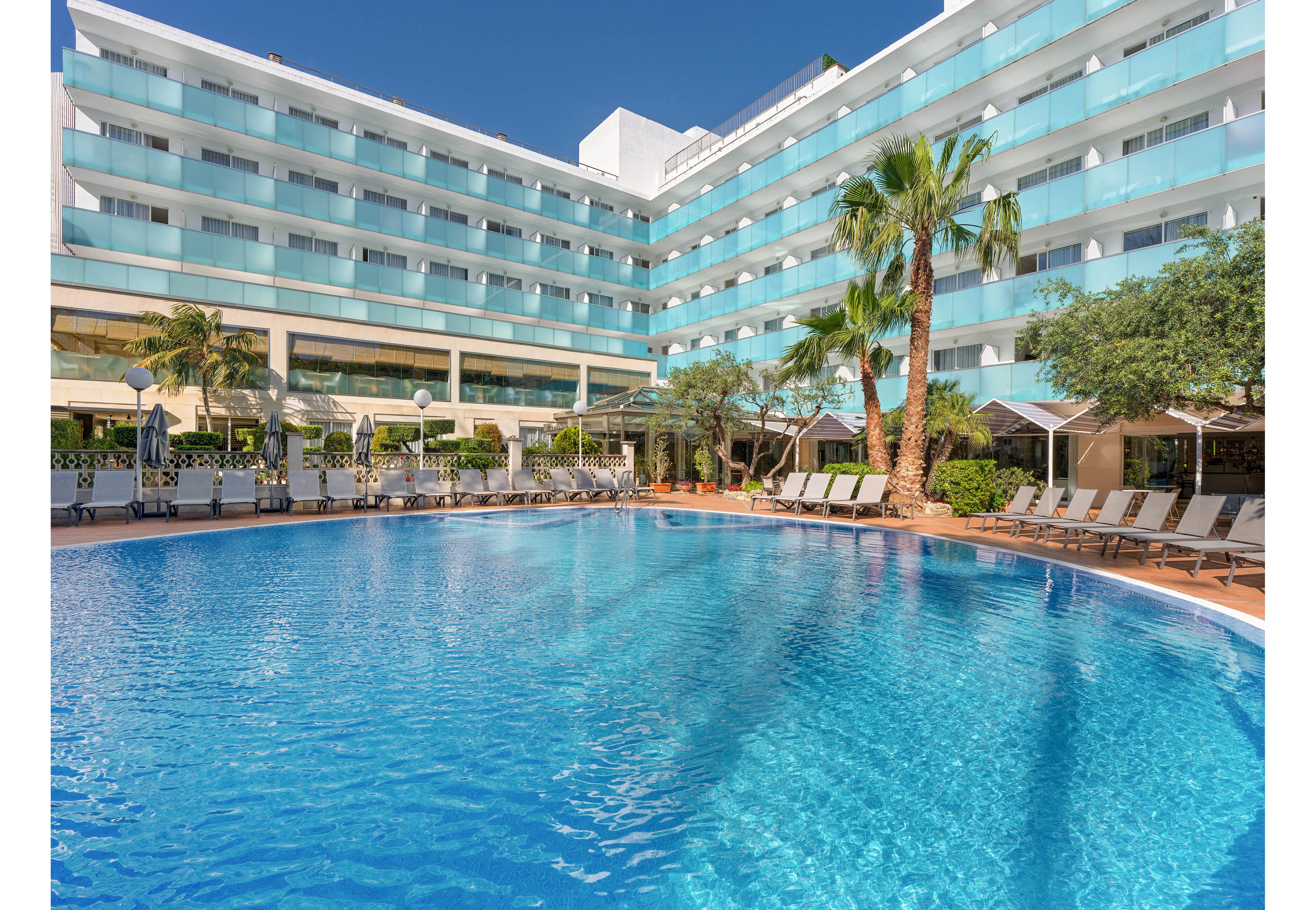 A large, modern hotel building overlooks a bright blue outdoor swimming pool. Multiple stories of rooms with glass balconies face the pool area, creating a resort-like atmosphere. Sun loungers line the pool deck, shaded by palm trees and greenery. The clear water and sunny sky suggest a warm, relaxing vacation setting.