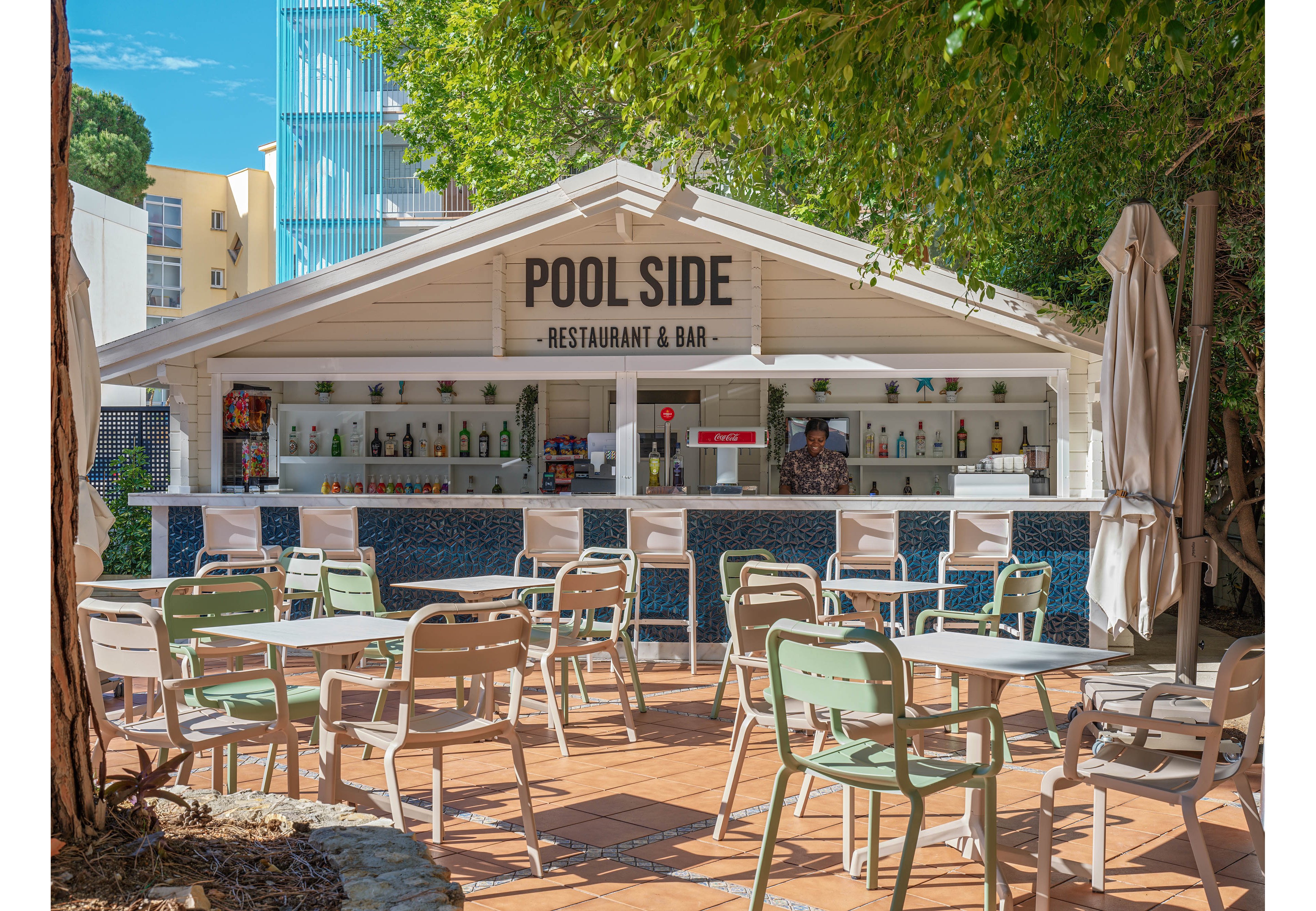 An open-air poolside restaurant and bar features a white wooden structure with a sign reading 'POOL SIDE RESTAURANT & BAR.' The bar counter is lined with various bottles and glassware, while metal and plastic chairs surround small tables on a tiled patio. Tall trees and greenery provide shade, and folded umbrellas stand near the seating area. Modern buildings rise in the background, suggesting a resort or hotel setting on a sunny day.