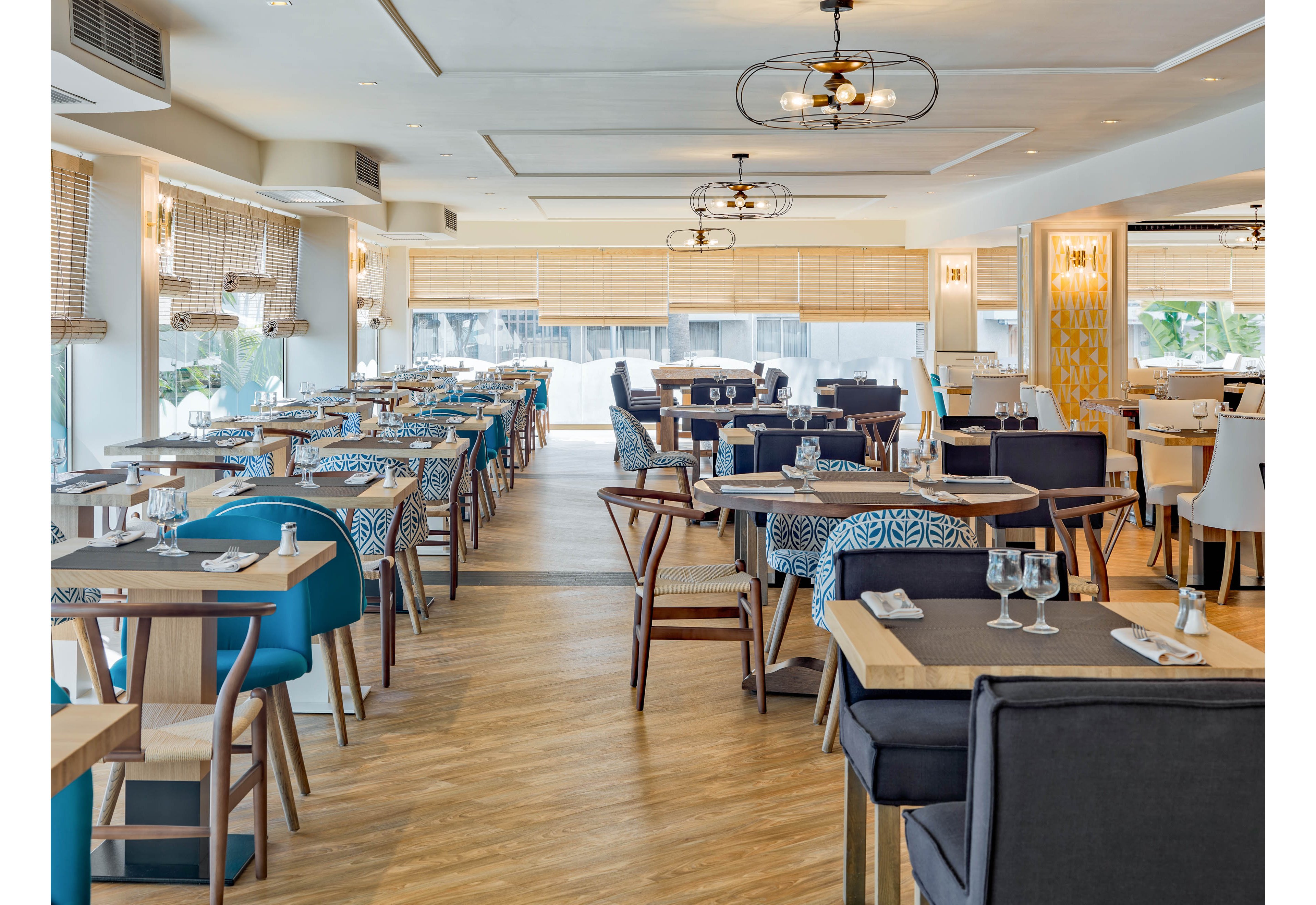 A contemporary restaurant dining room features neatly arranged wooden tables and upholstered chairs. The space is bright and airy, with large windows, light-colored walls, and woven blinds allowing natural light to fill the room. Table settings with glassware and napkins are prepared for guests, while patterned cushions and teal accents add visual interest. Ceiling lights and wooden flooring contribute to the warm, inviting atmosphere of the interior.