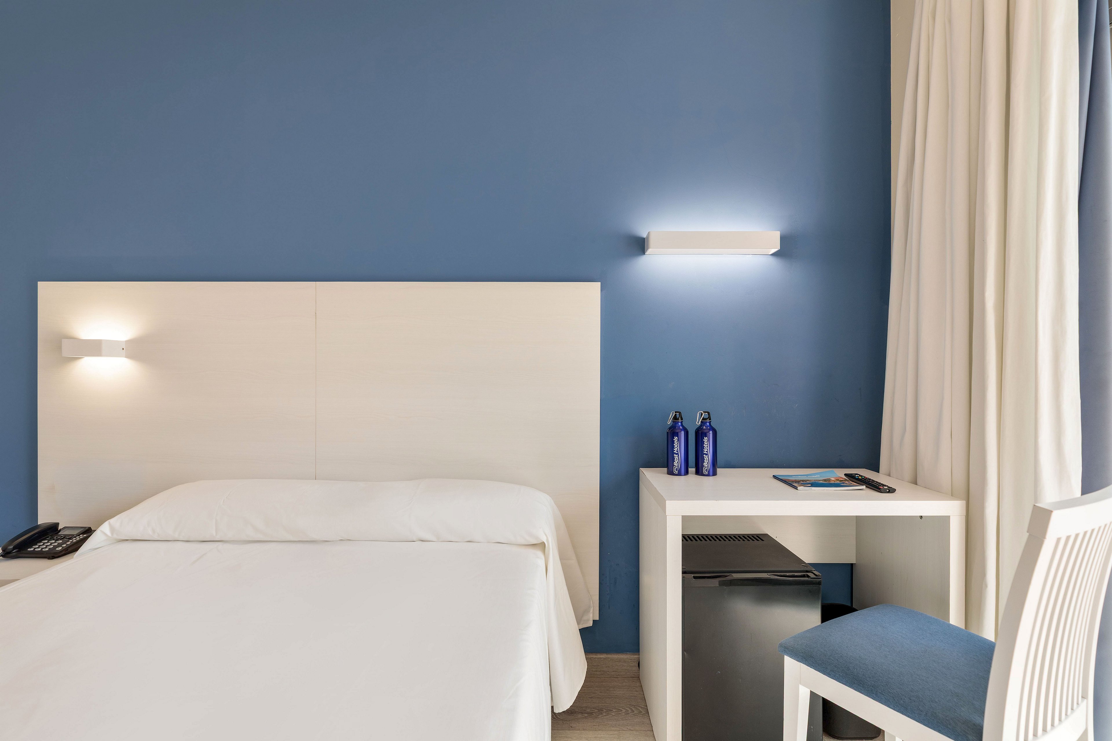 A bright, minimalist hotel room features a neatly made single bed with a light-colored headboard against a blue accent wall. A small white desk with a chair sits beside the bed, holding two blue bottles and a notepad. Beneath the desk is a compact mini fridge, while wall-mounted lights and sheer curtains add to the clean, contemporary look. Natural light filters through the window, creating a calm and inviting atmosphere.