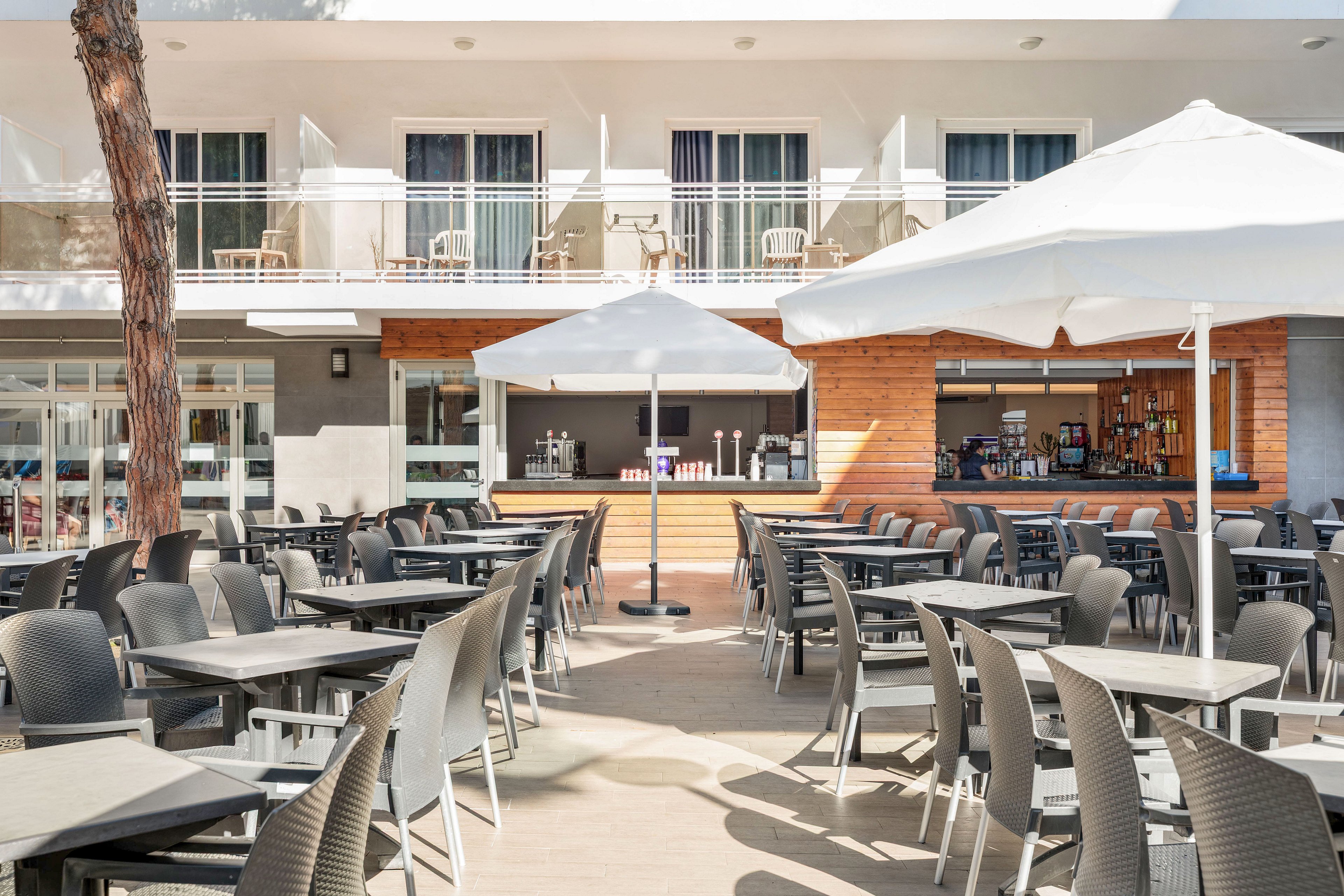 Bright outdoor terrace bar area with rows of empty tables and chairs arranged neatly under large white umbrellas. The setting appears to be part of a modern hotel or resort, with balconies and glass doors visible in the background. The bar counter features various bottles and drink dispensers, suggesting a casual dining or refreshment space. Overall, the scene is clean, sunlit, and ready to welcome guests.