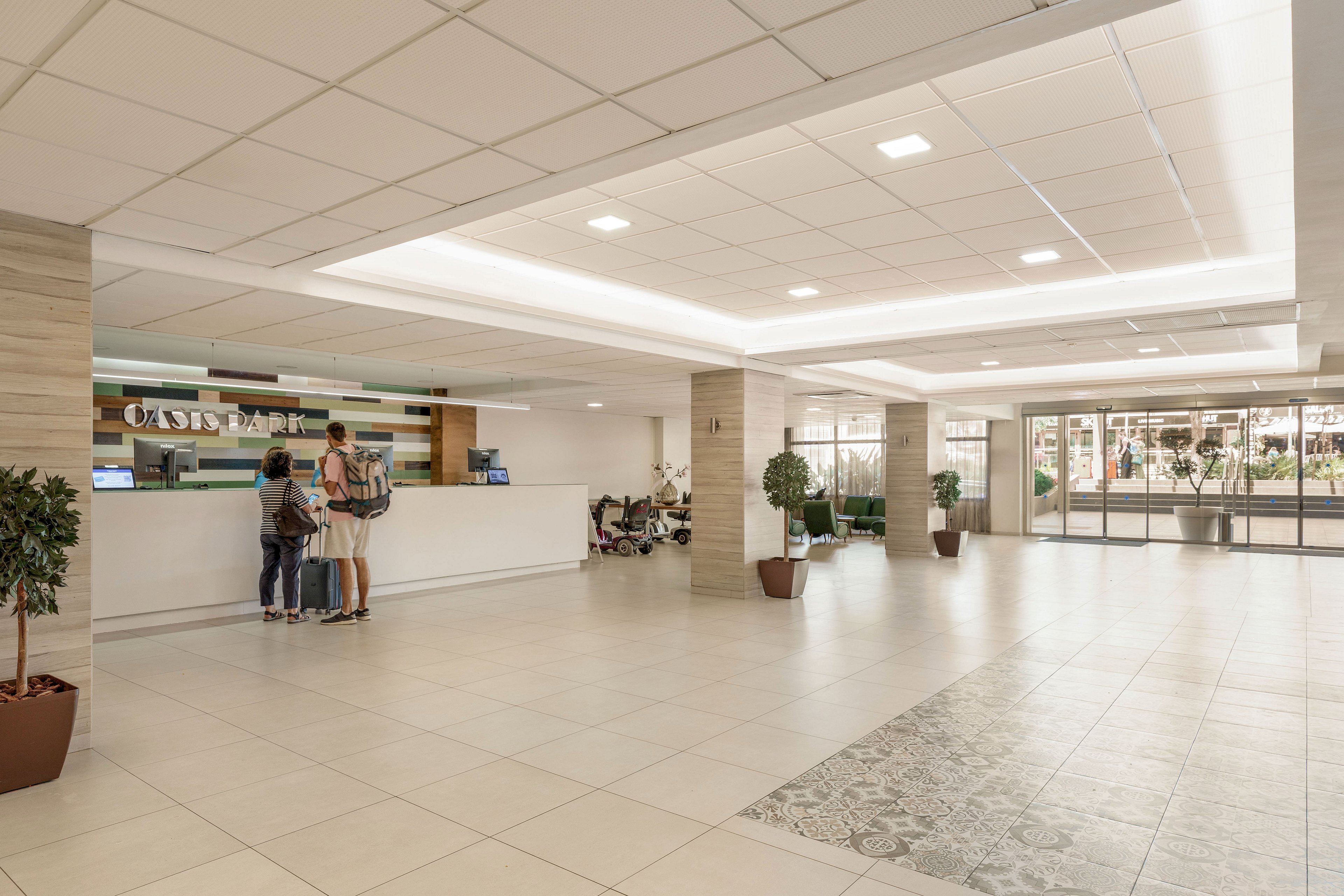 A spacious, modern lobby features a bright reception desk where three people are speaking with a staff member. The area has large tiled floors, recessed ceiling lights, and neutral-colored walls with wood accents. Potted plants and seating areas add warmth to the contemporary interior. Large glass doors at the far end open to an outdoor area, allowing natural light to fill the space.