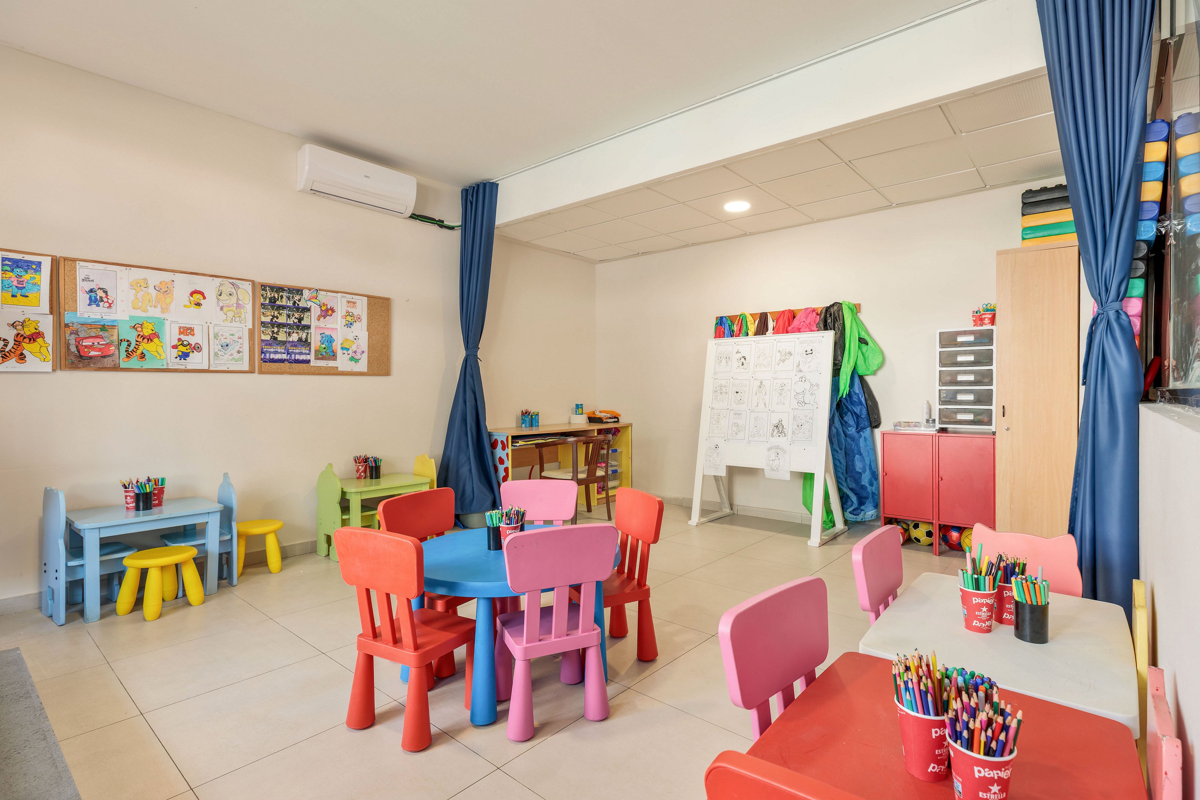 Brightly colored child-sized tables and chairs fill a preschool classroom, each topped with cups of crayons and markers. The room features educational posters and children’s drawings on the wall, along with a whiteboard easel and storage cabinets. Blue curtains frame the space, and the neutral floor and ceiling contrast with the vivid reds, blues, yellows, and greens of the furniture. The overall setting suggests a creative, organized environment for early childhood learning and play.