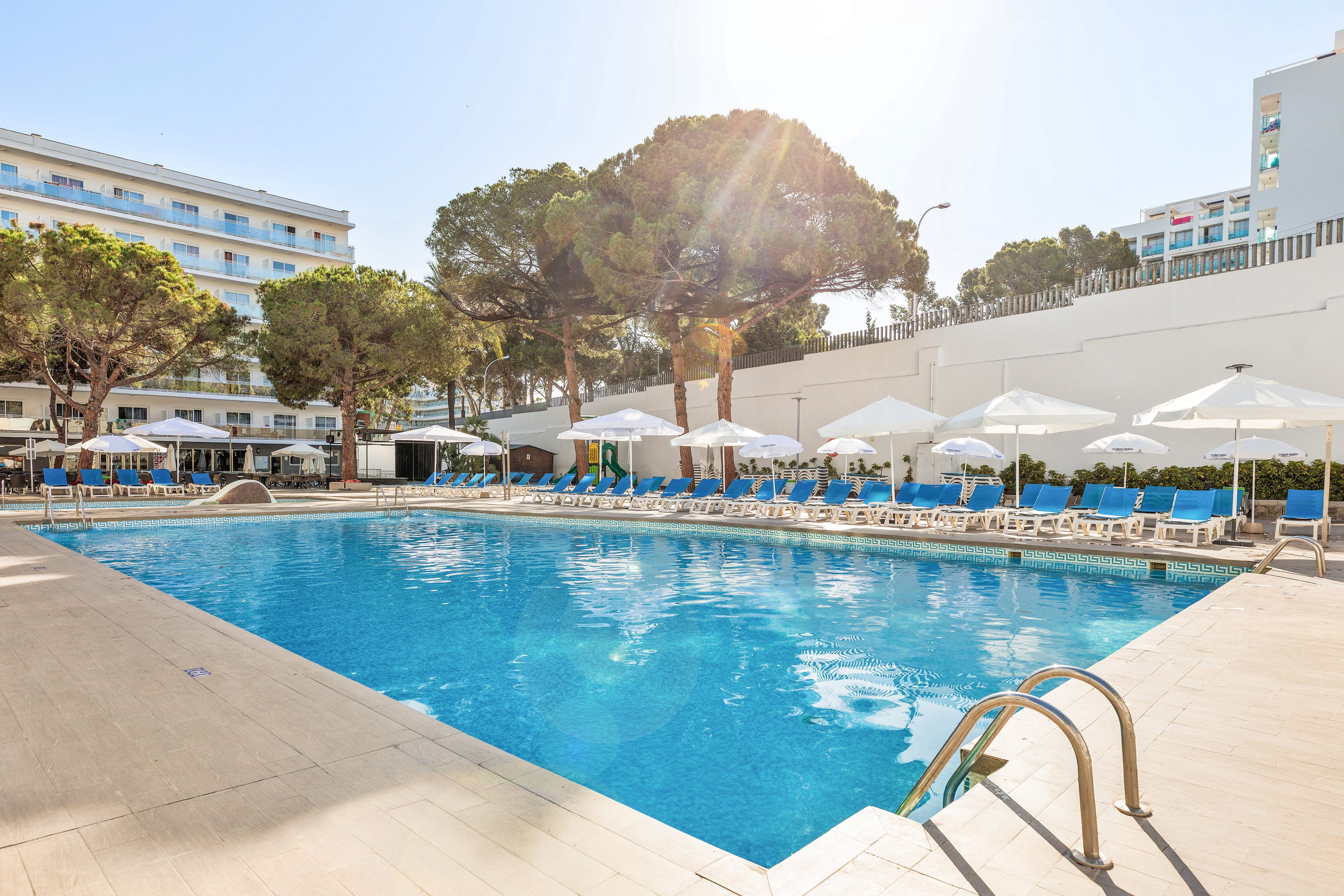 Large blue outdoor swimming pool is surrounded by sun loungers and white umbrellas at a modern hotel. Tall trees provide natural shade along the far side of the pool area. The bright sunlight and clear sky create a warm, inviting vacation atmosphere. Nearby multi-story hotel buildings frame the leisure space, suggesting a resort or holiday destination.