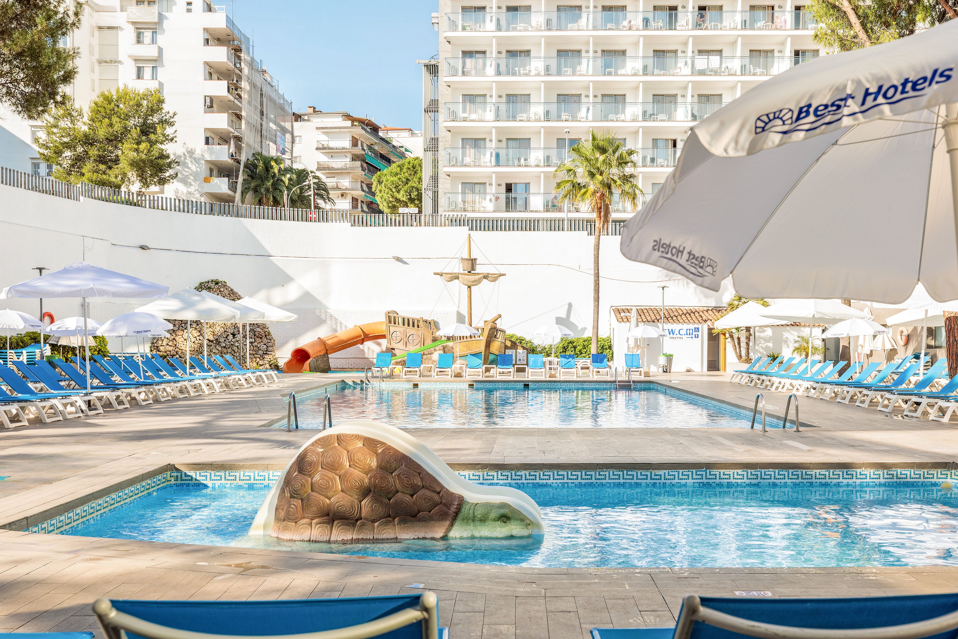 Bright daytime view of a hotel outdoor swimming pool surrounded by blue sun loungers and umbrellas. A multi-story hotel building rises in the background, with balconies overlooking the pool. A small waterslide and playful fountain feature are visible near the far end of the pool. White umbrellas with clearly readable "Best Hotels" text provide shade around the deck, creating a relaxed vacation atmosphere.