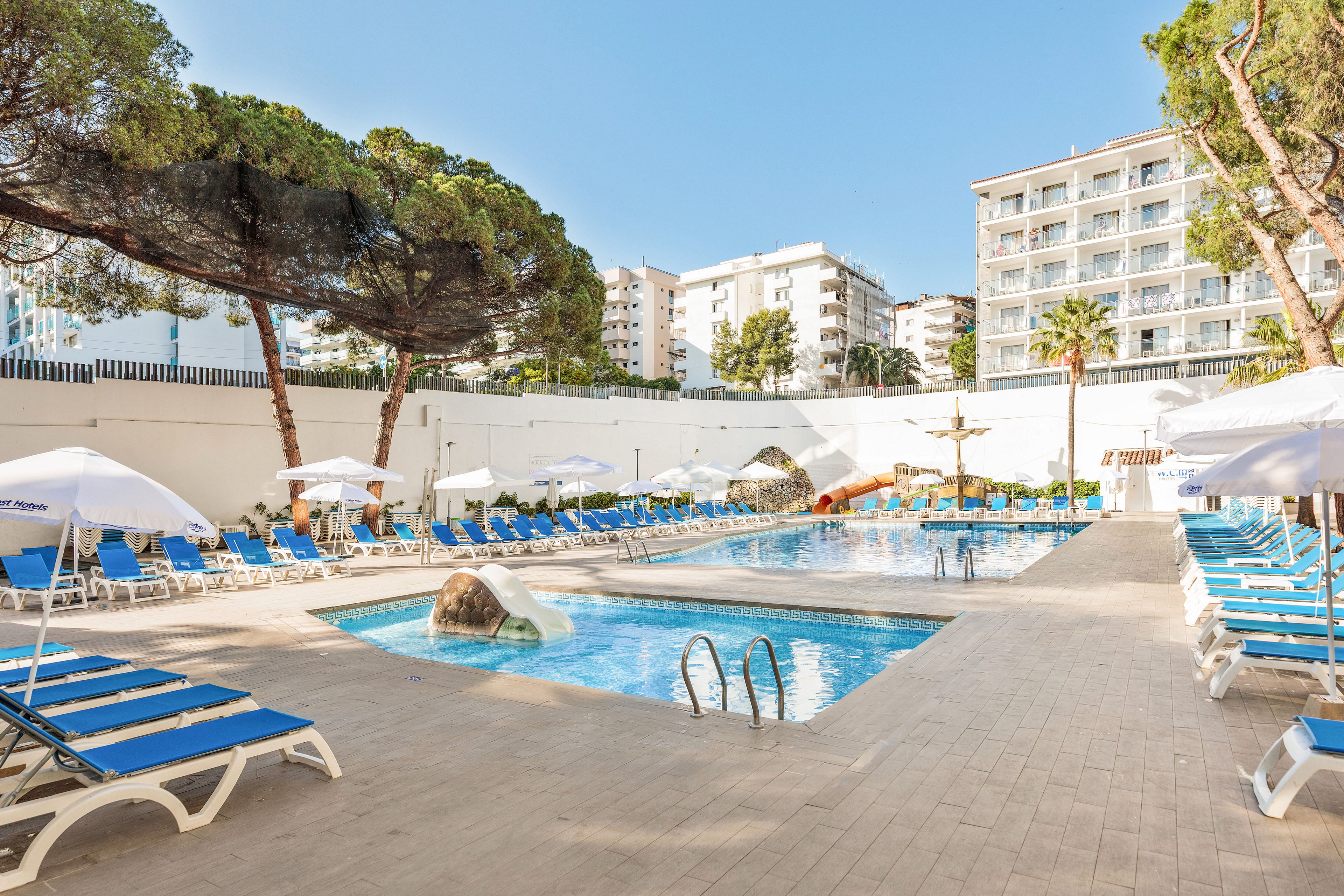 Bright outdoor hotel pool area with blue sun loungers and white umbrellas arranged around a rectangular swimming pool. A small rock feature and metal ladders are visible in the clear water, with a fountain structure at the far end. Tall trees provide partial shade, and a multi-story hotel building rises in the background under a clear blue sky. The scene suggests a relaxed vacation atmosphere in a warm, sunny destination.