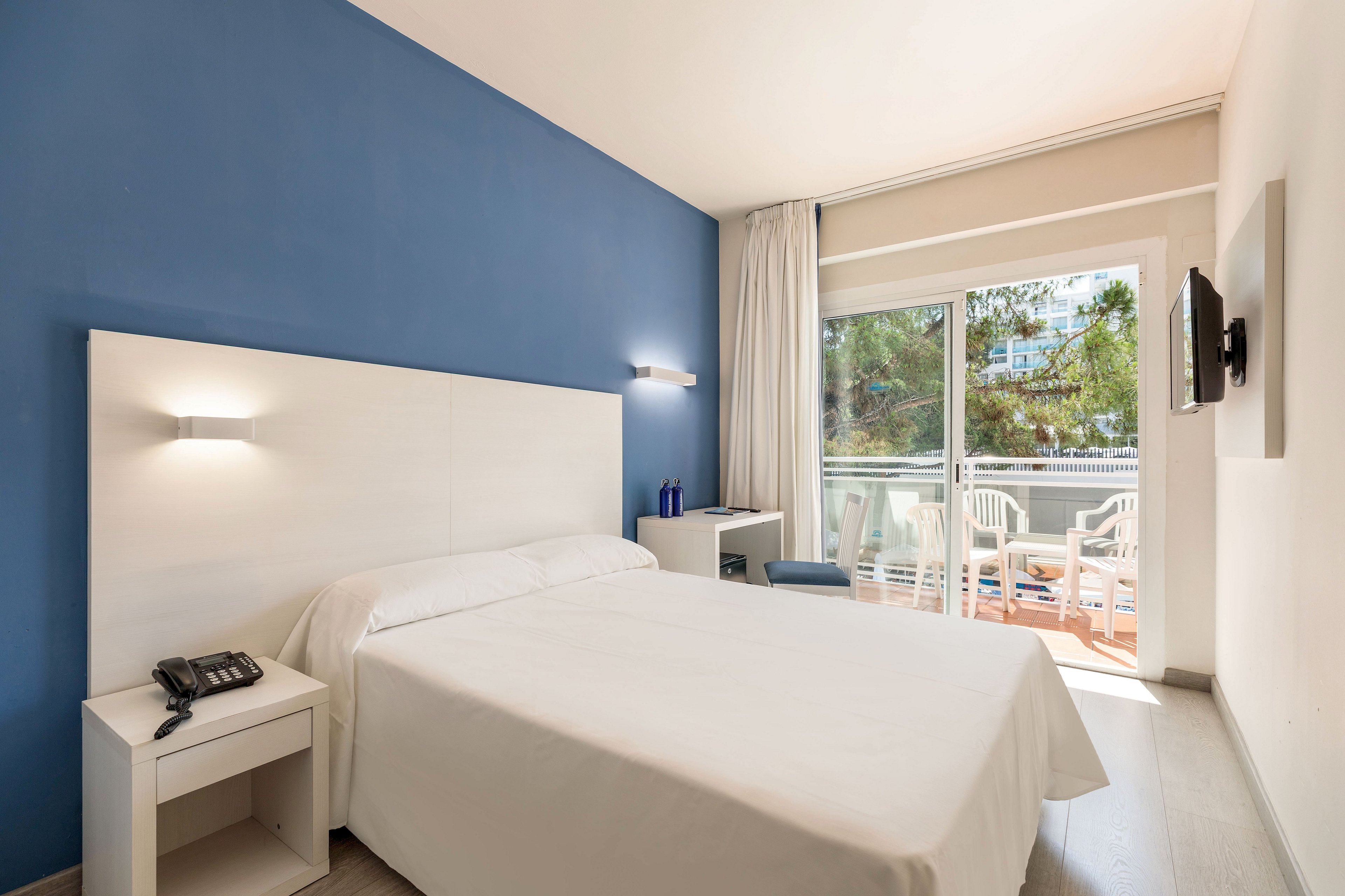 A bright modern hotel room features a neatly made double bed against a blue accent wall. Minimalist white furniture, including a bedside table, desk, and wall-mounted TV, create a clean contemporary look. Large sliding glass doors open onto a balcony with white chairs and a view of trees and nearby buildings. Natural daylight fills the space, giving the room an airy and inviting atmosphere.
